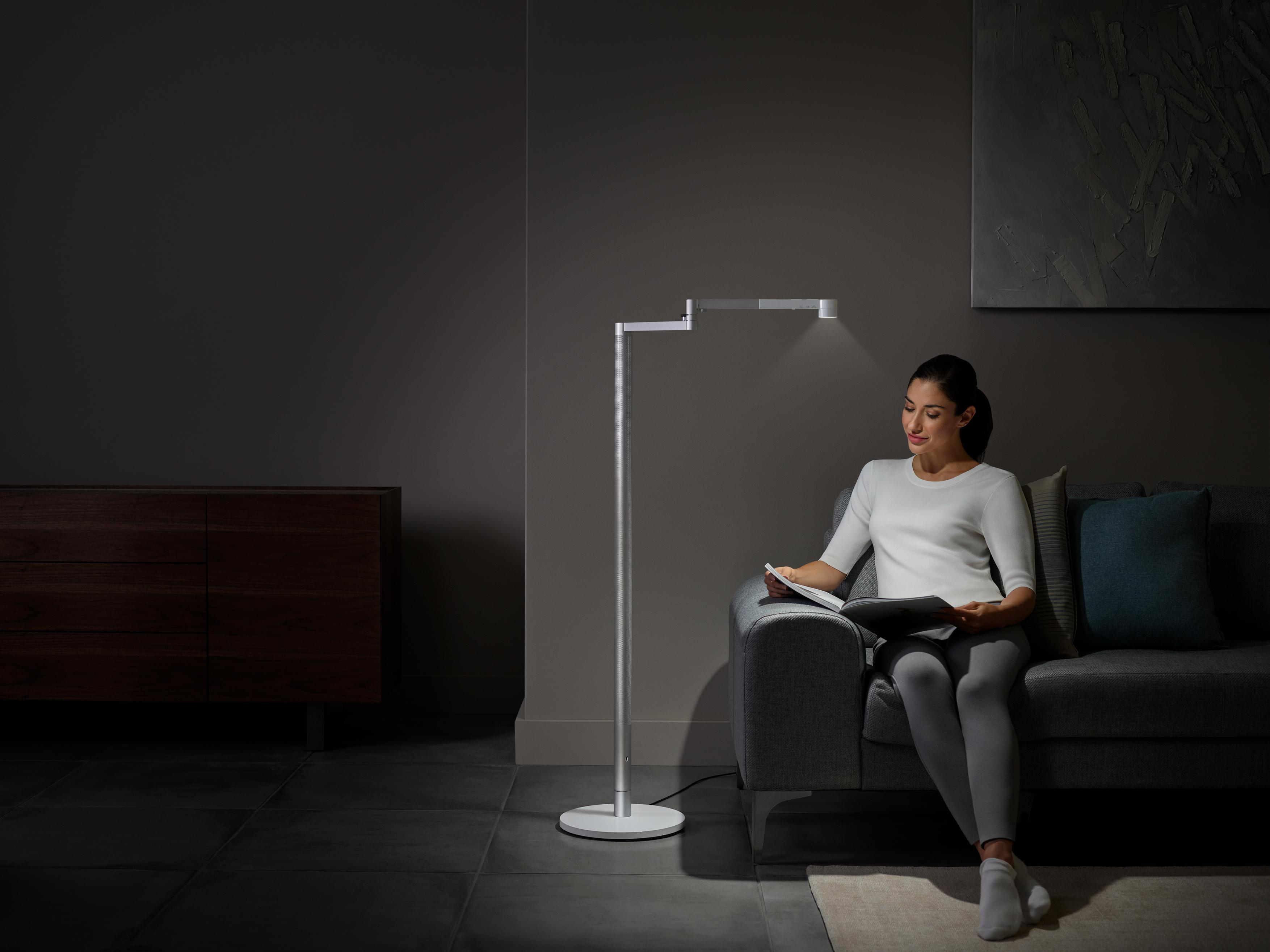 Frau liest unter einer modernen Stehlampe auf einem Sofa in einem schwach beleuchteten Raum.