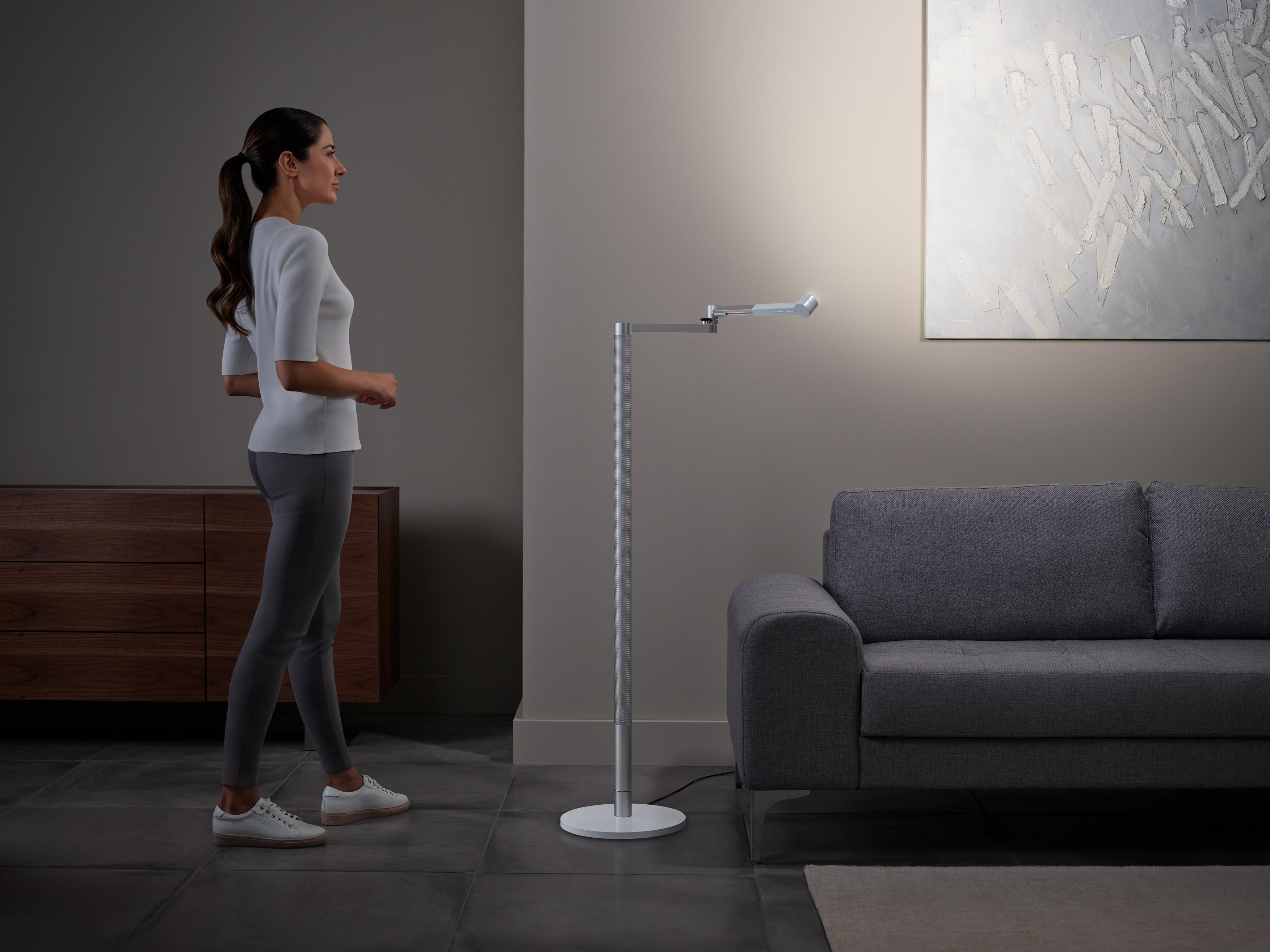 Eine Frau steht neben einer modernen Stehlampe. Graues Sofa und abstrakte Kunst im Hintergrund.