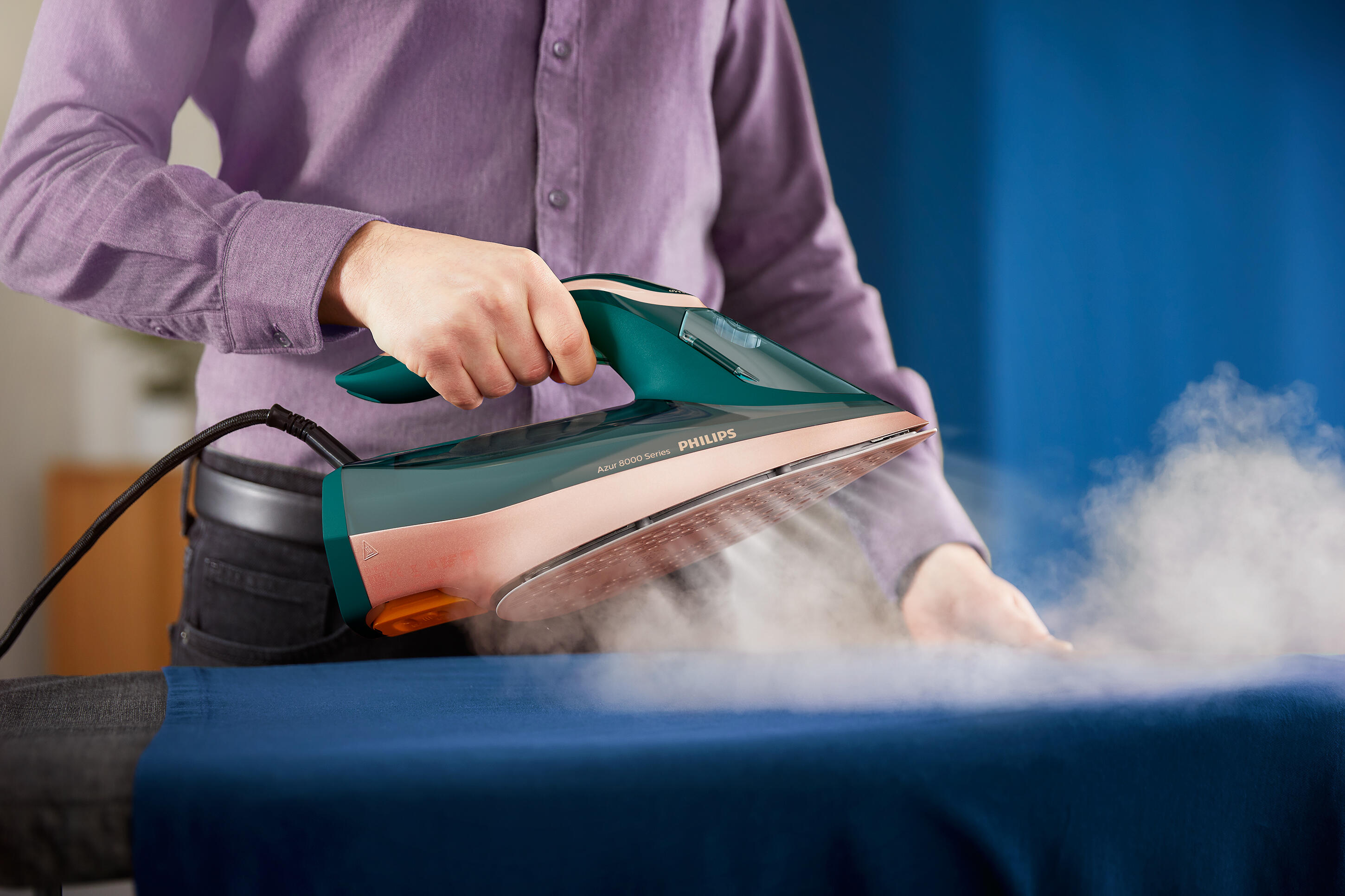 Una persona planchando tela azul con una plancha verde y rosa que emite vapor.