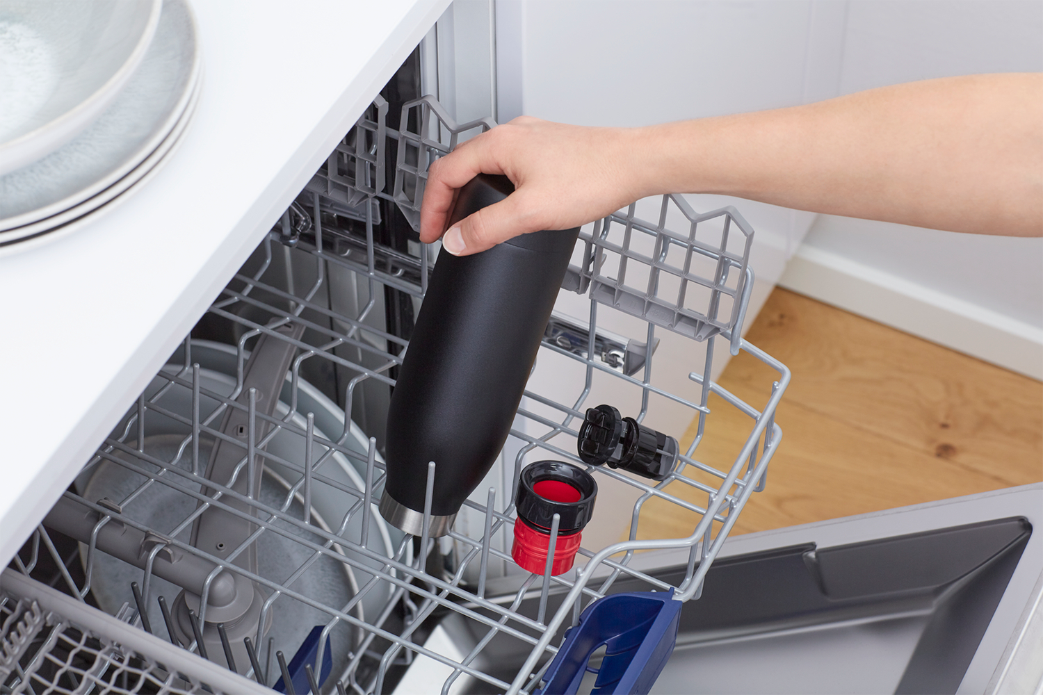 A hand places a black water bottle into a dishwasher. Other items are visible in the rack.