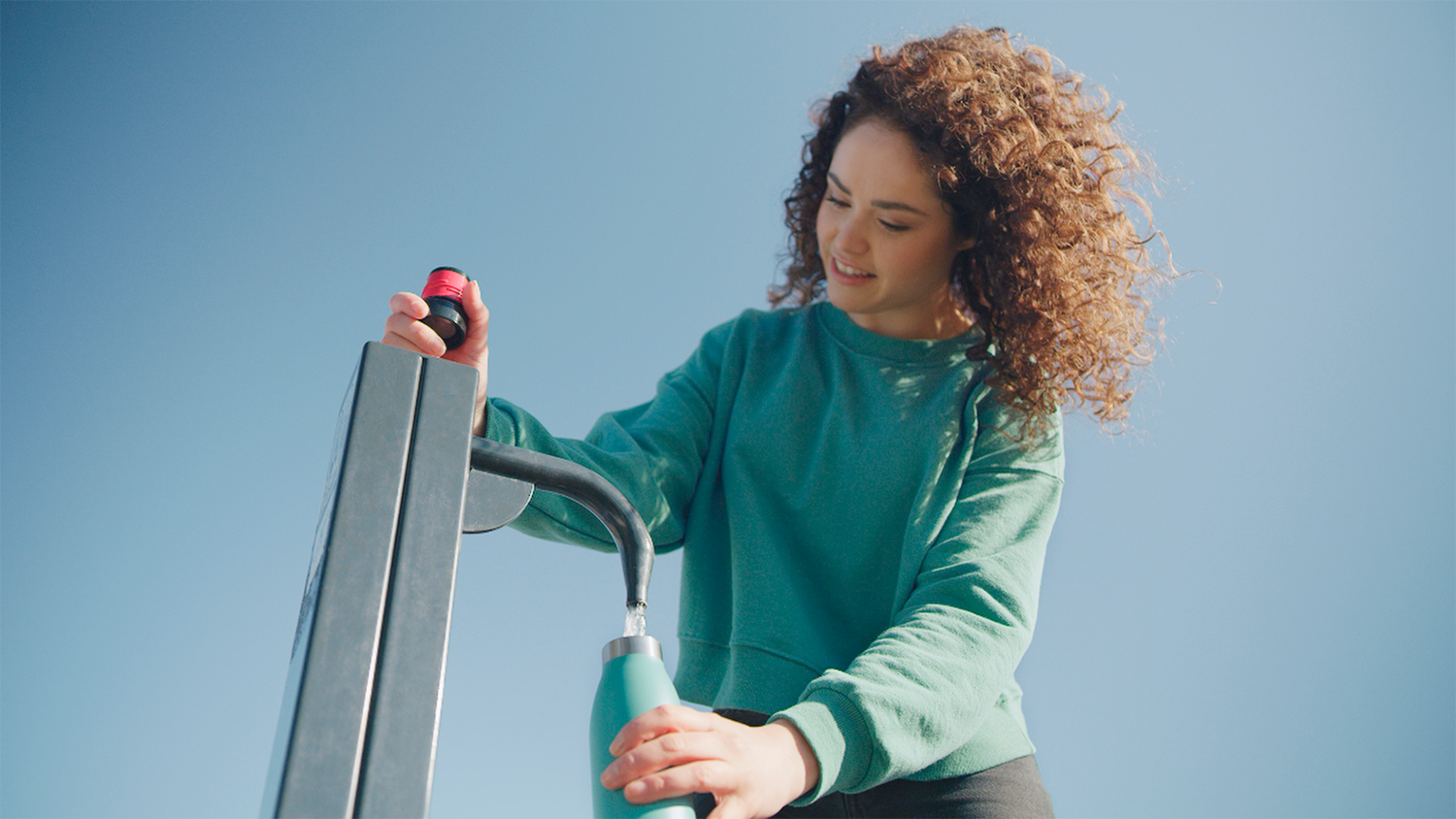 Vrouw vult een waterfles met een kraan, tegen een blauwe lucht.