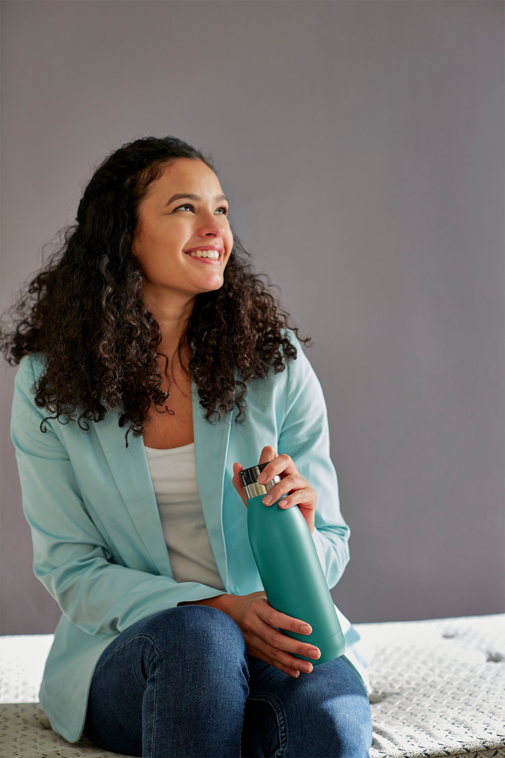 Lachende vrouw met krullend haar, blauwe jas, houdt een teal waterfles vast, kijkt omhoog.