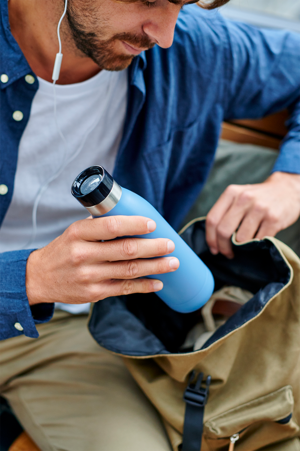 Man putting a blue water bottle into a backpack, wearing a blue shirt and khaki pants.