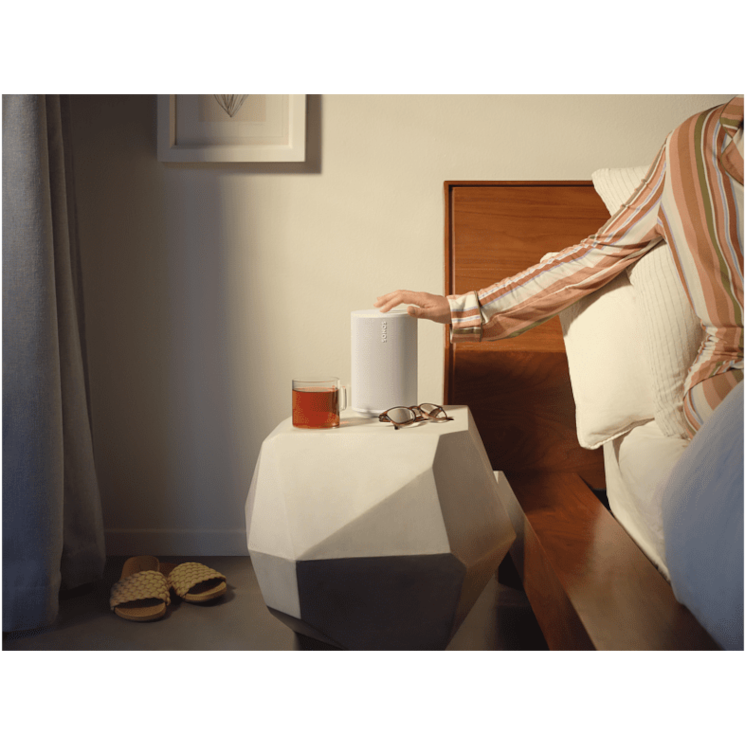 Een arm reikt naar een witte speaker op een stenen tafel. Een kopje en bril zijn aanwezig.