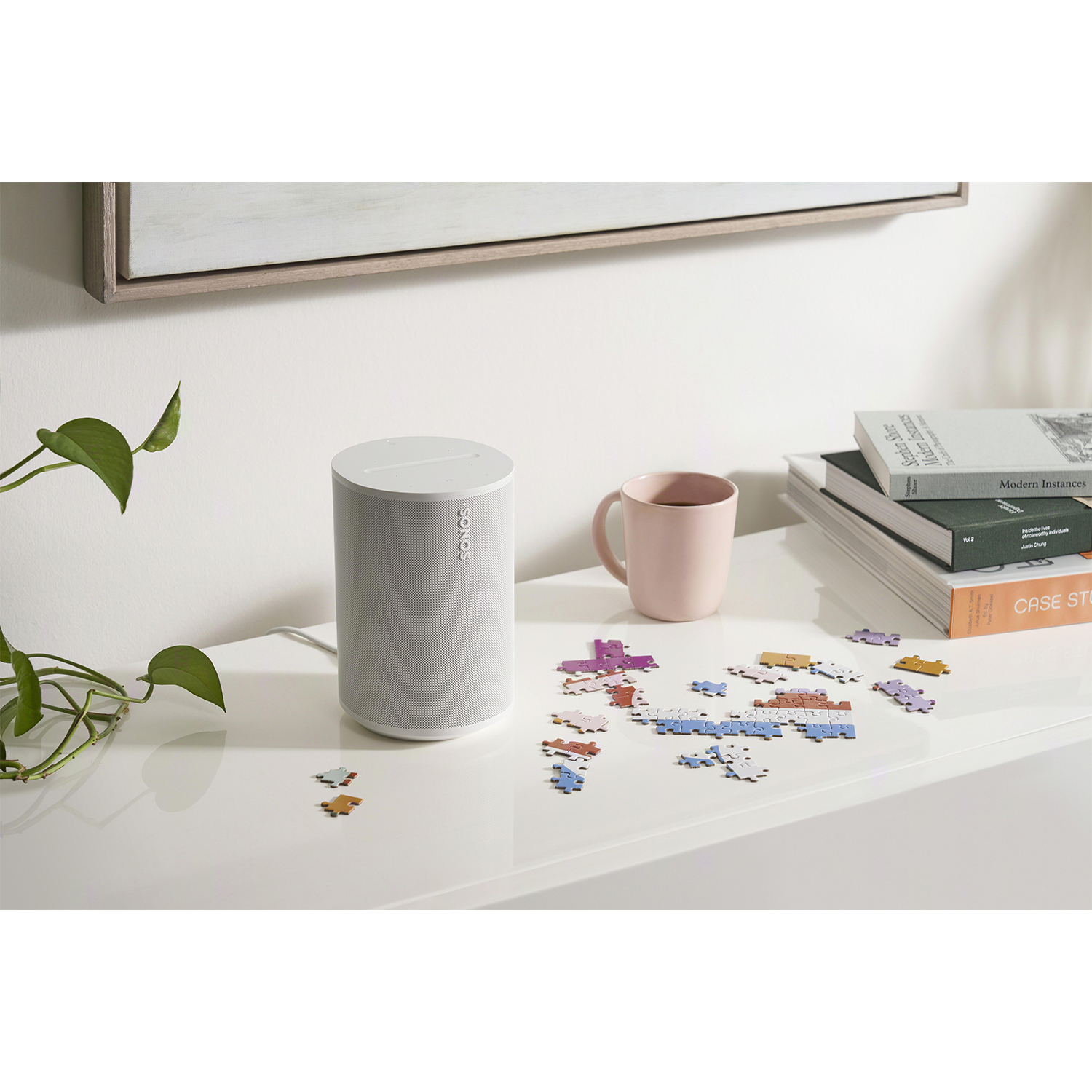 Un altoparlante bianco, una tazza rosa, pezzi di puzzle, libri e un quadro su un tavolo bianco.
