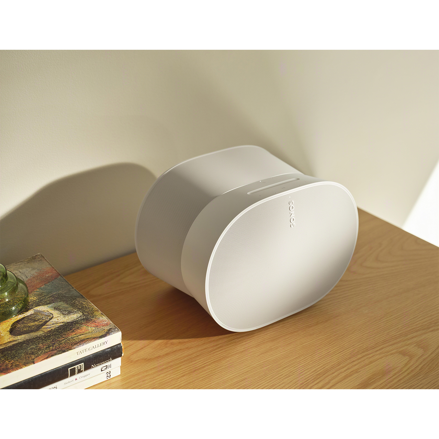 Altoparlante Sonos bianco su un tavolo di legno con libri e un vaso verde.
