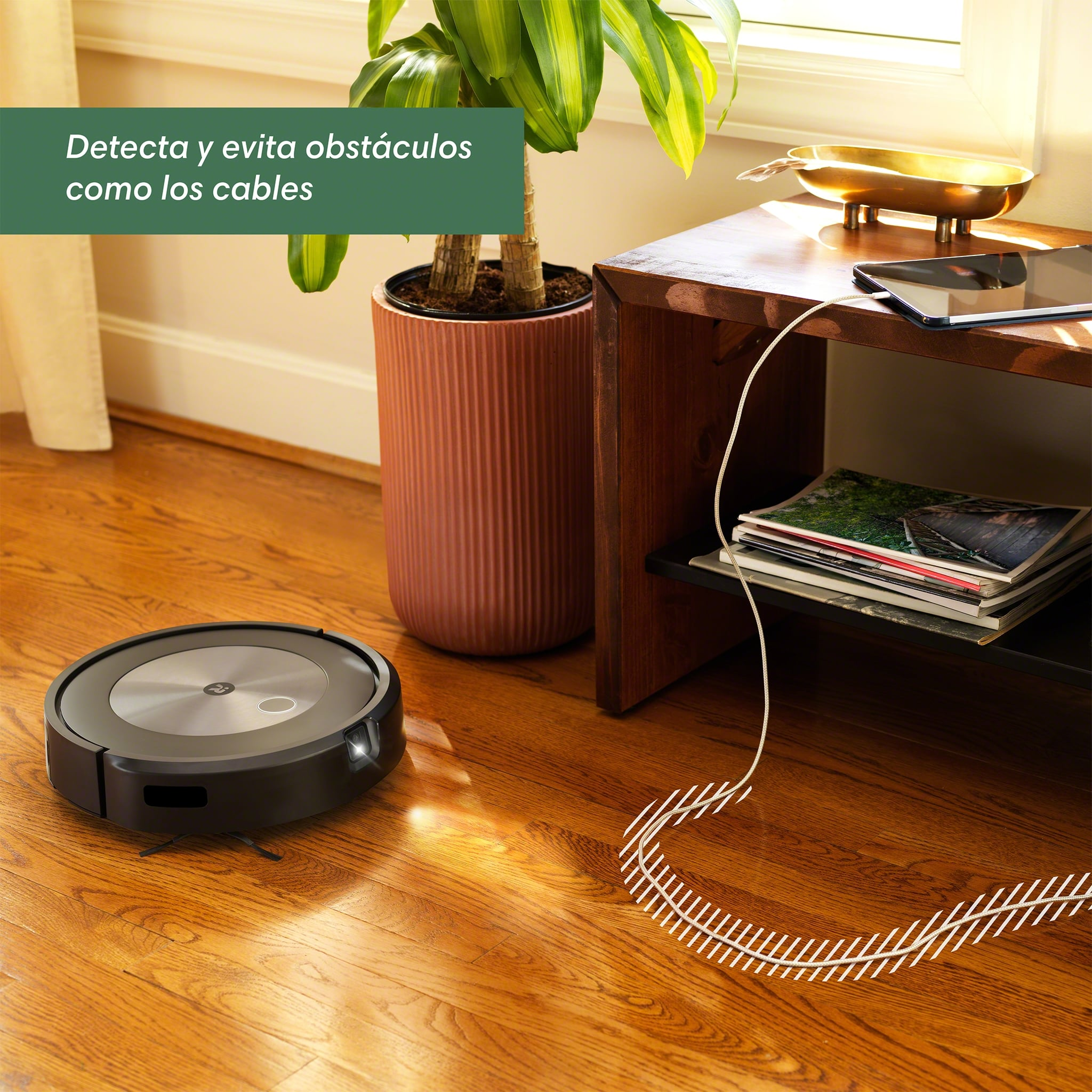 Aspiradora robot negra en piso de madera, cerca de planta y mesa.