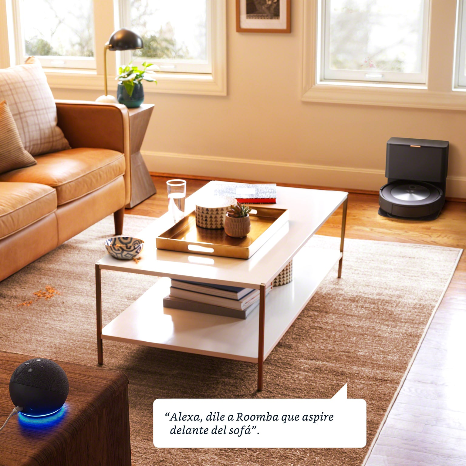 Salón con mesa de centro blanca, un dispositivo Alexa y un Roomba. Burbuja de texto: Alexa, dile a Roomba.