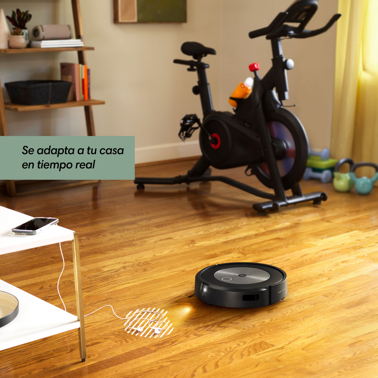 Una aspiradora robot negra en suelo de madera. Una bicicleta estática y otros objetos en el fondo.