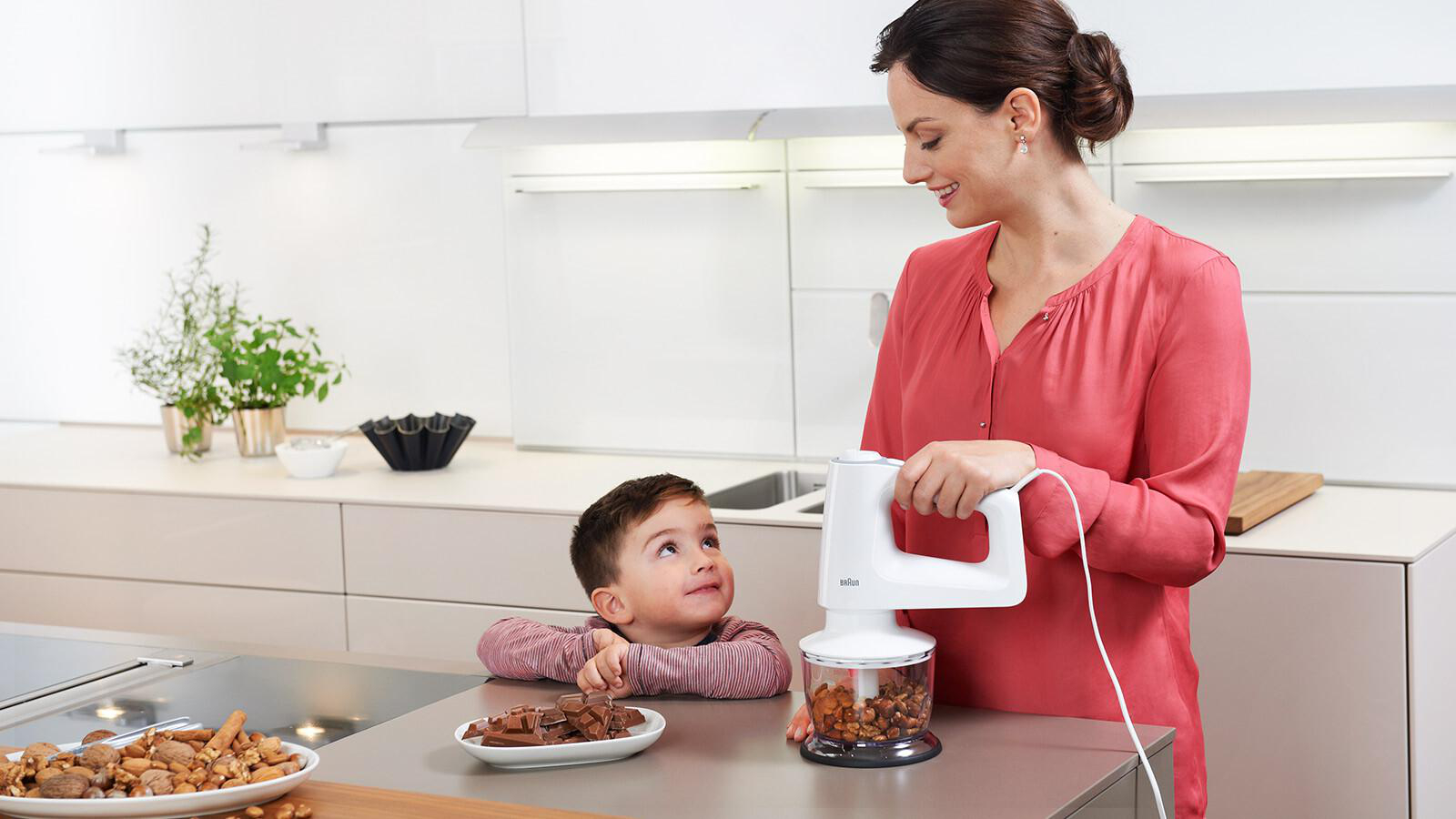 Frau und Kind bereiten Essen mit einem weißen Handmixer zu. Nüsse und Schokolade liegen auf der Theke.