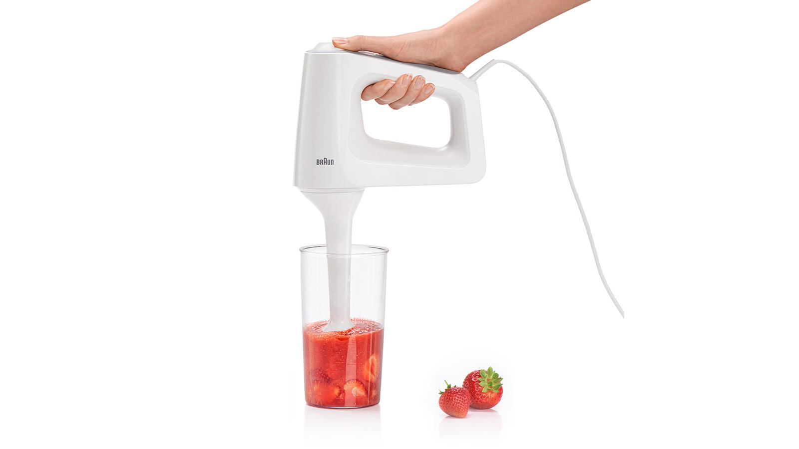 Hand hält weißen Braun Stabmixer in einem Glas mit pürierten Erdbeeren.