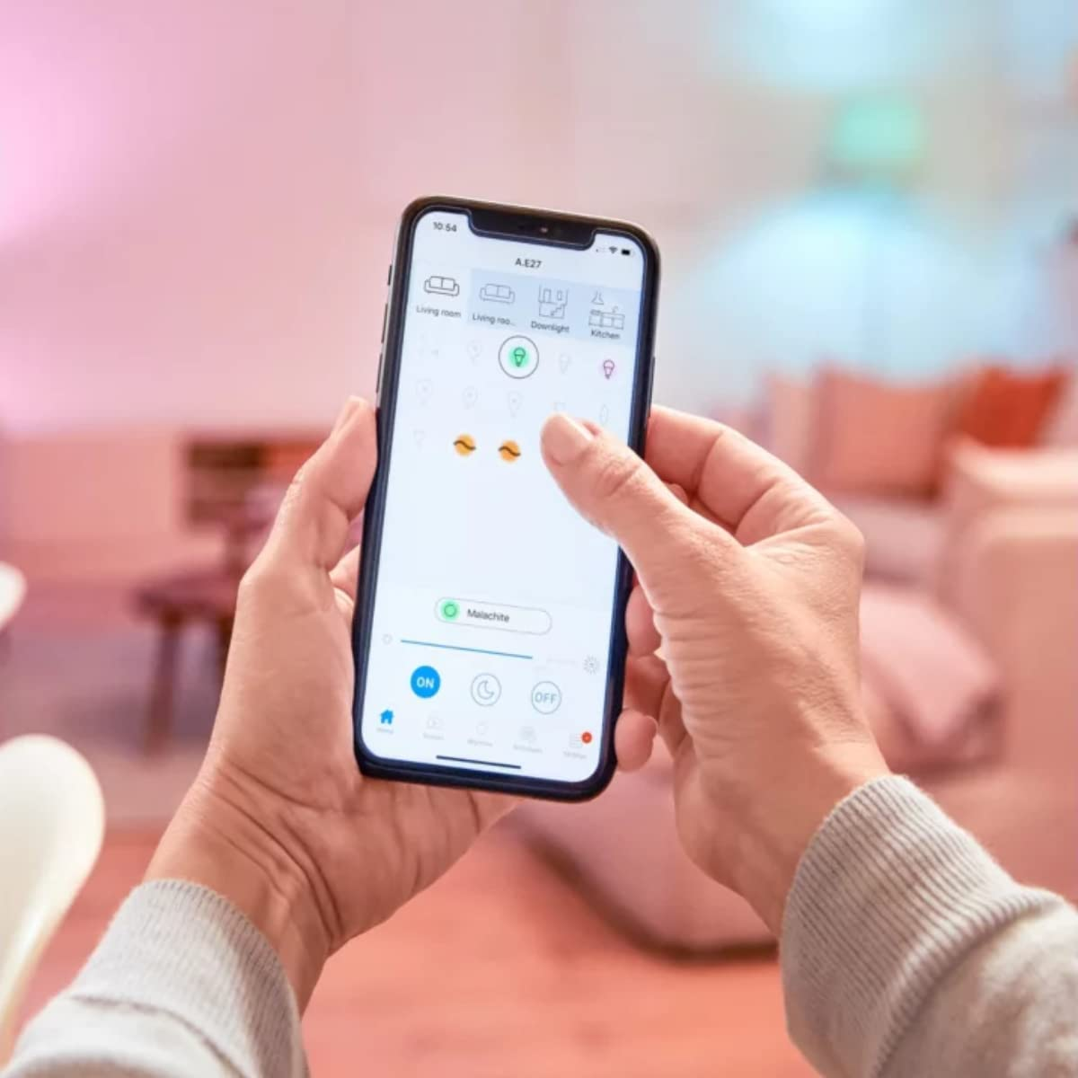 Una persona sostiene un teléfono inteligente que muestra una interfaz de aplicación para el hogar inteligente. La aplicación controla luces y otros dispositivos.