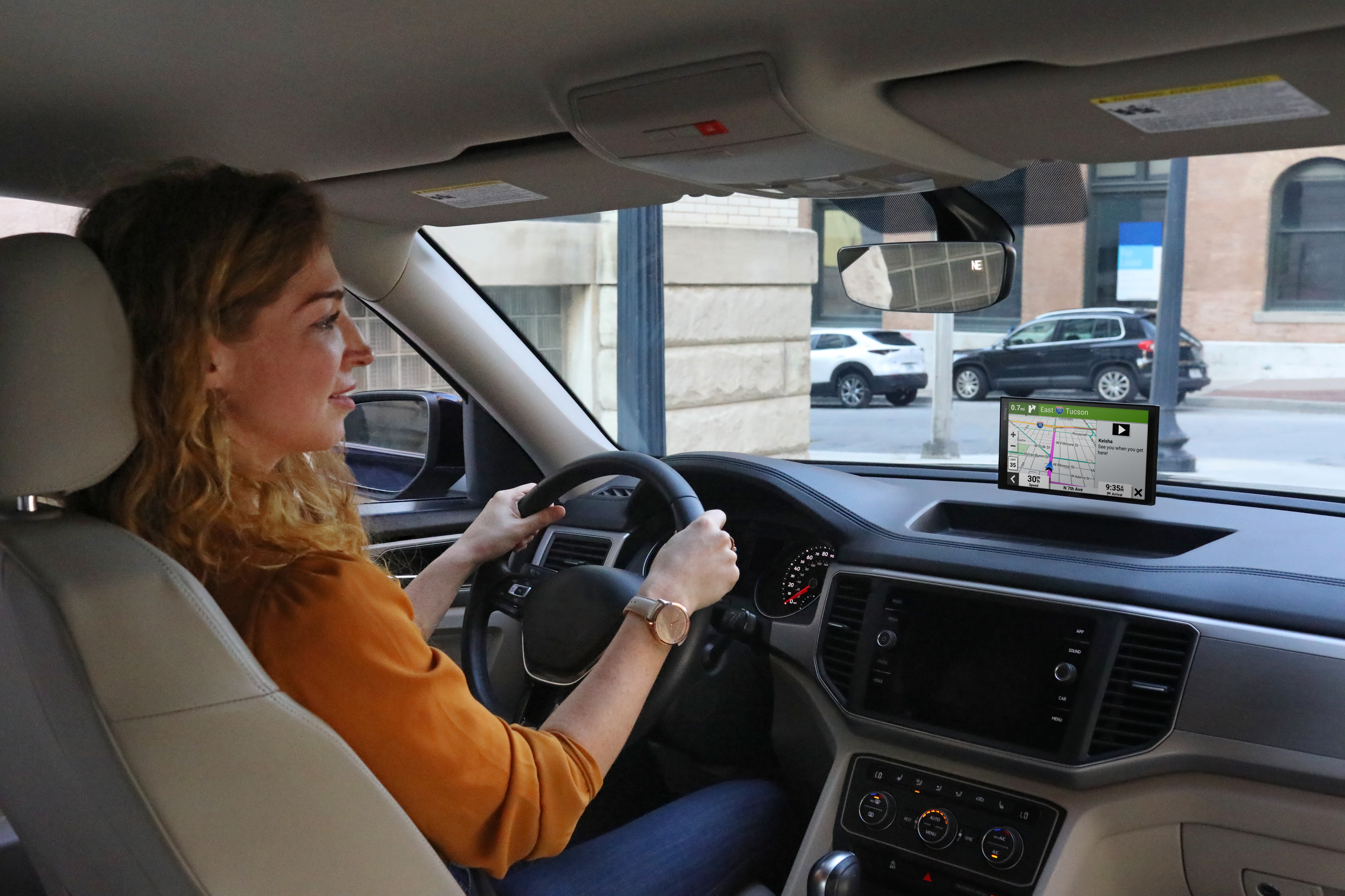 Donna alla guida, mani sul volante, sguardo in avanti. GPS acceso. Edifici e auto sullo sfondo.