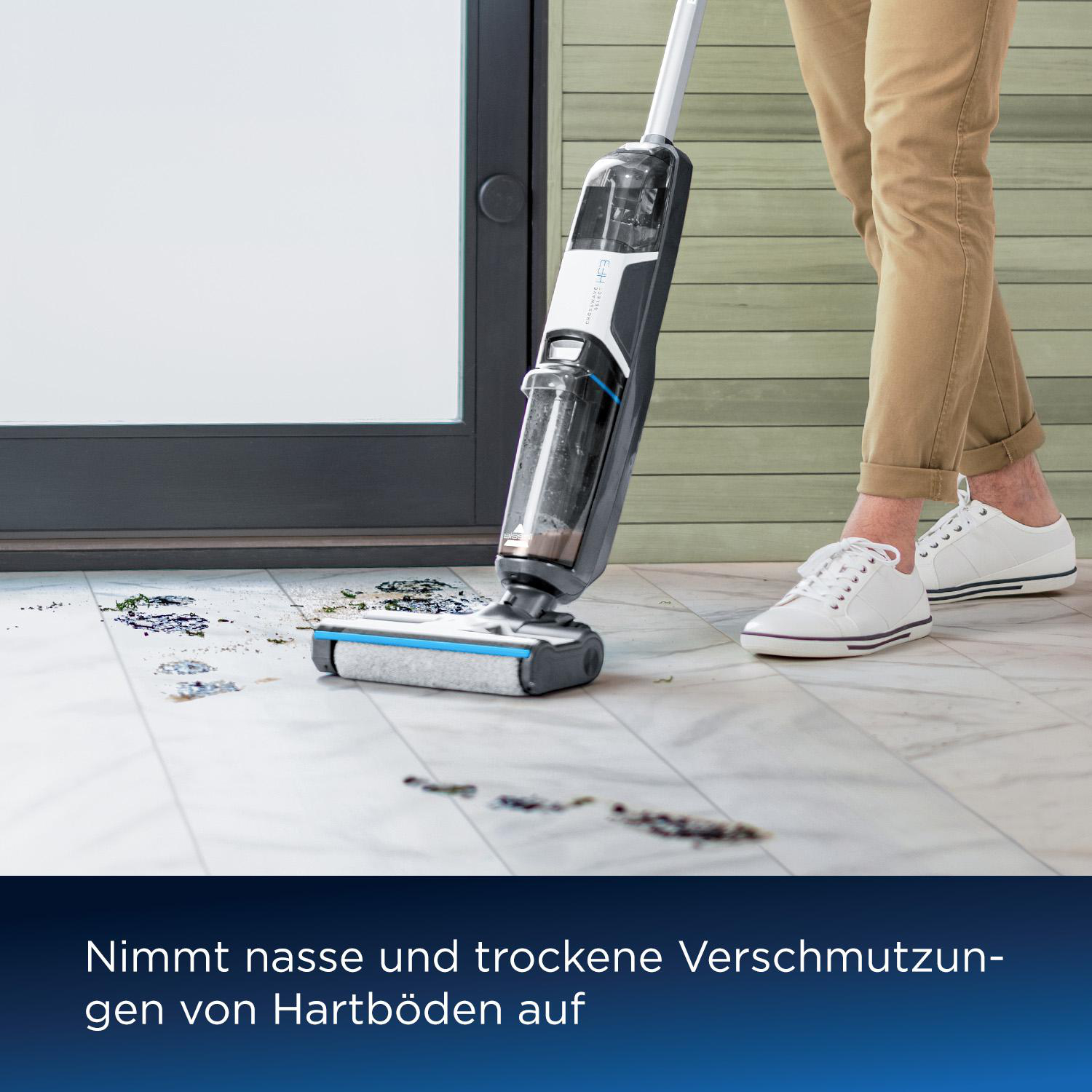 Eine Person benutzt einen Staubsauger auf einem schmutzigen Boden. Der Staubsauger ist schwarz und weiß. Die Person trägt eine beige Hose und weiße Schuhe.