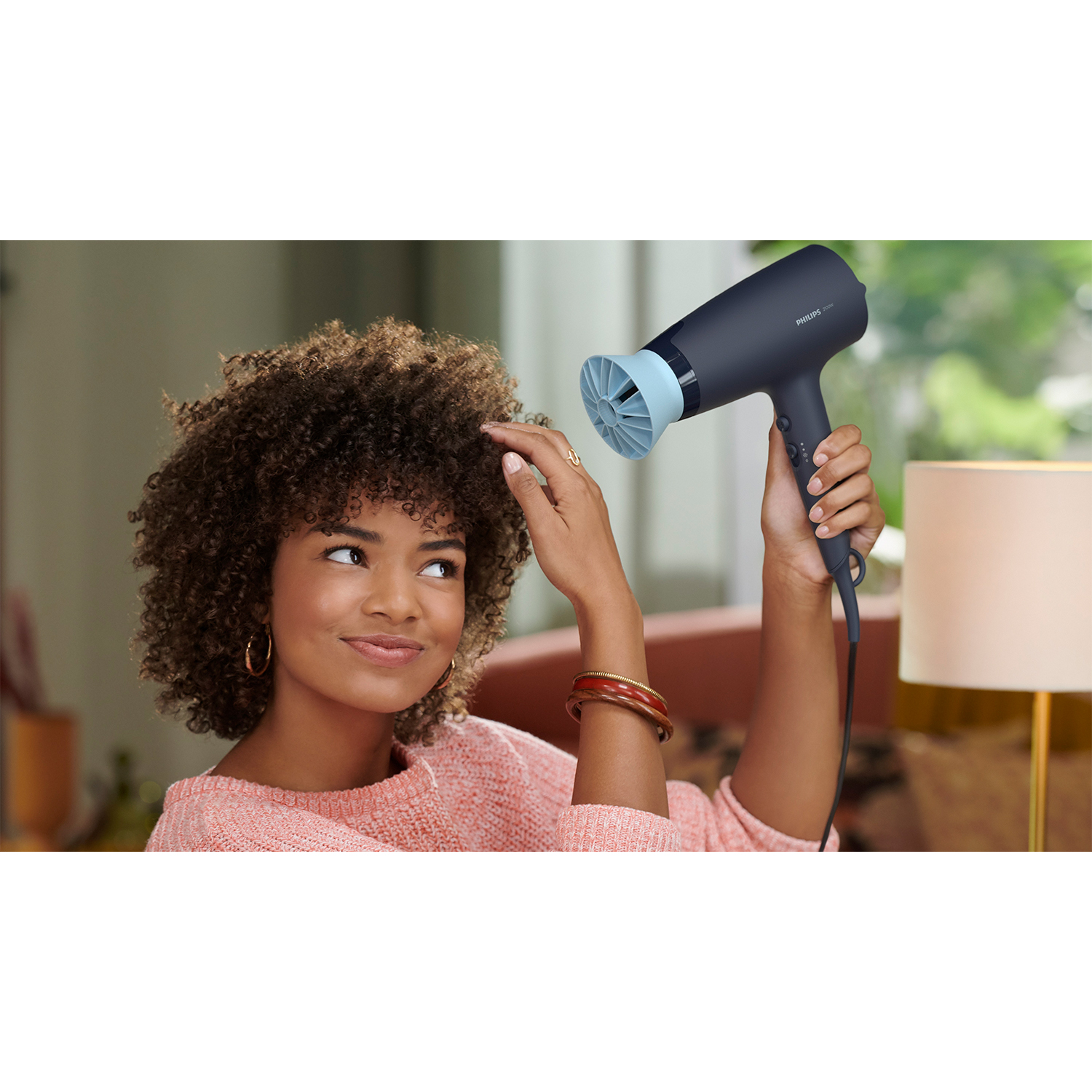 Una donna con i capelli ricci usa un asciugacapelli nero e blu. Sta guardando in alto, sorridendo.