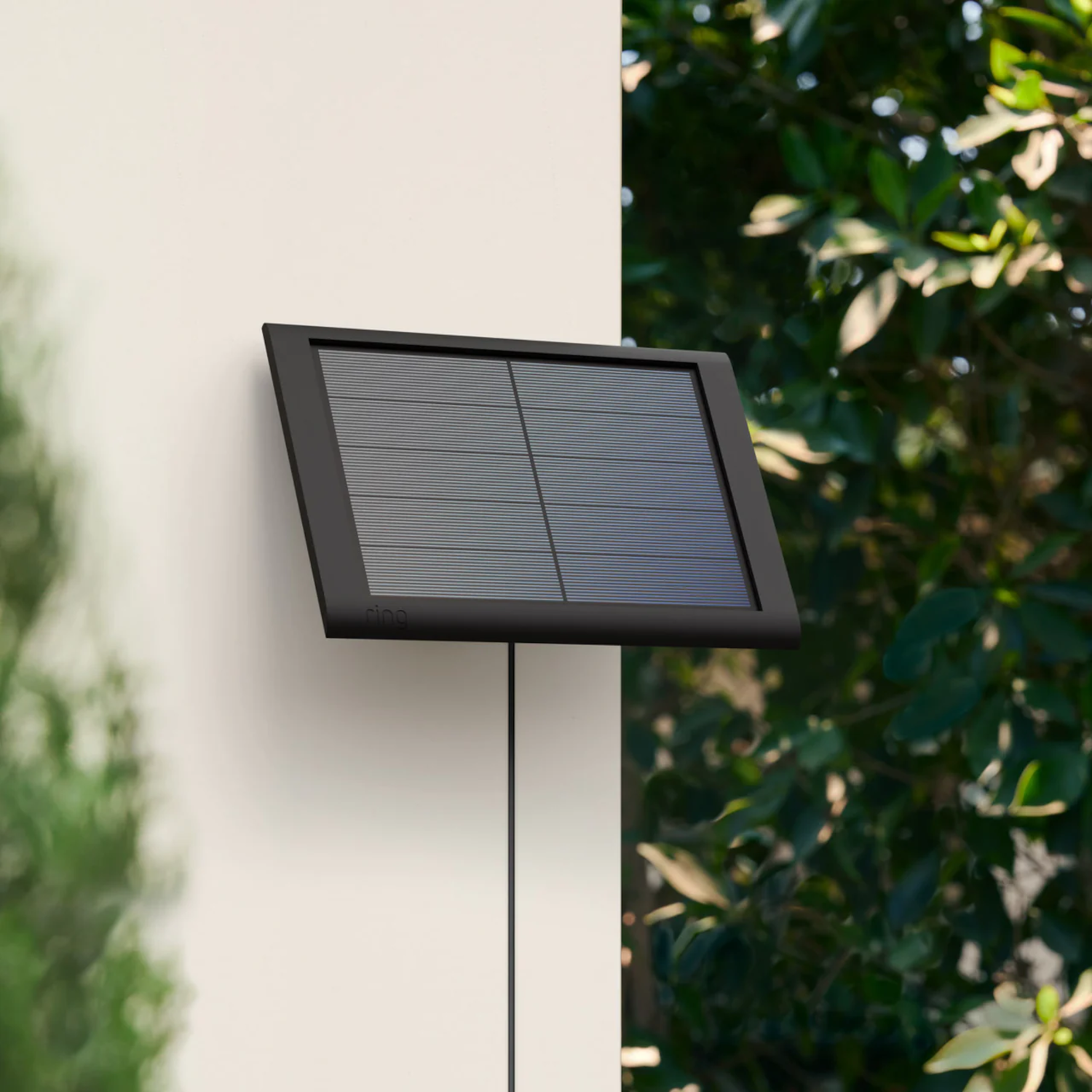 Black solar panel mounted on a white wall with green foliage in the background.