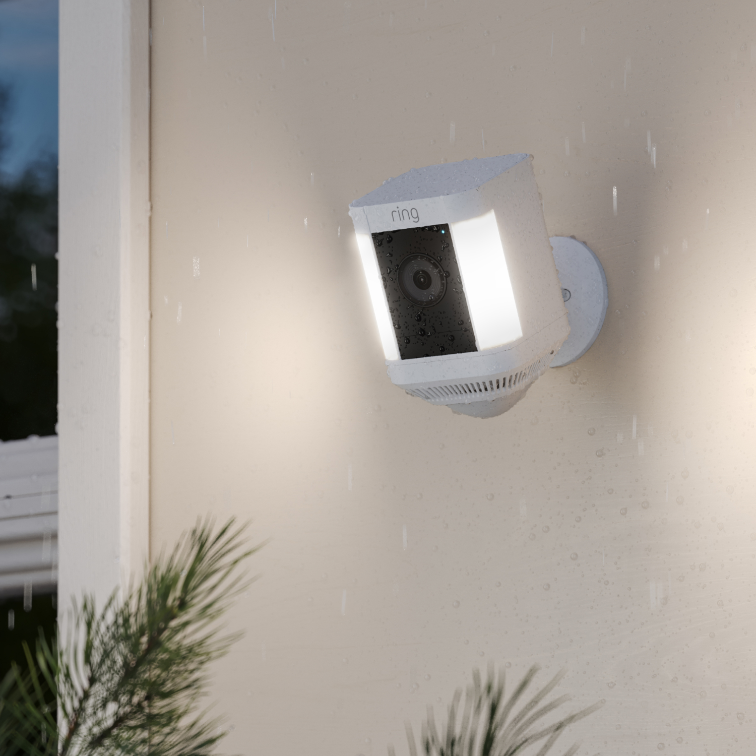 White Ring security camera mounted on a beige wall. It's raining. Green foliage in the corner.