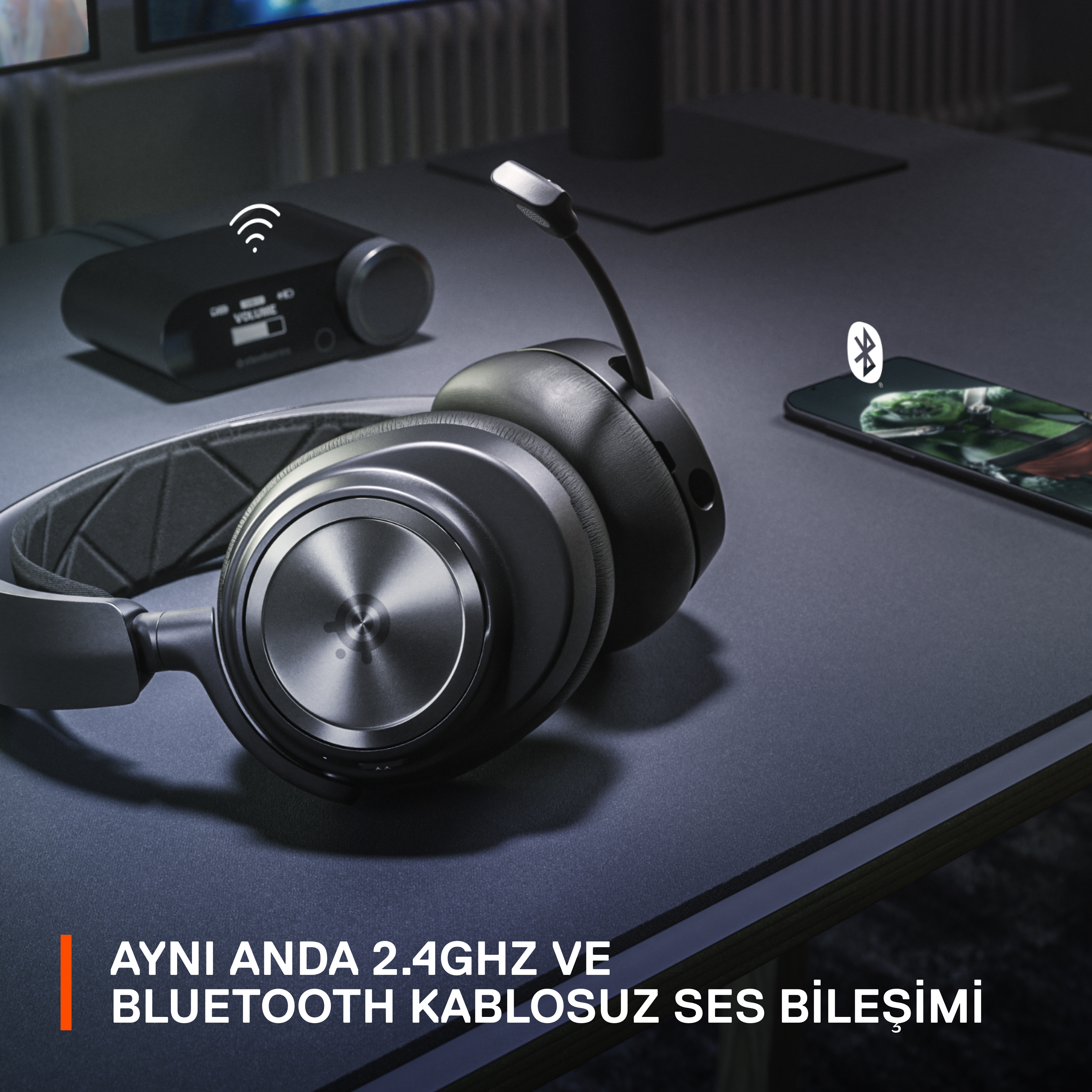 Siyah oyun kulaklığı, mikrofonlu, kablosuz, koyu renkli bir masa üzerinde. Metin: AYNI ANDA 2.4GHZ VE BLUETOOTH KABLOSUZ SES.