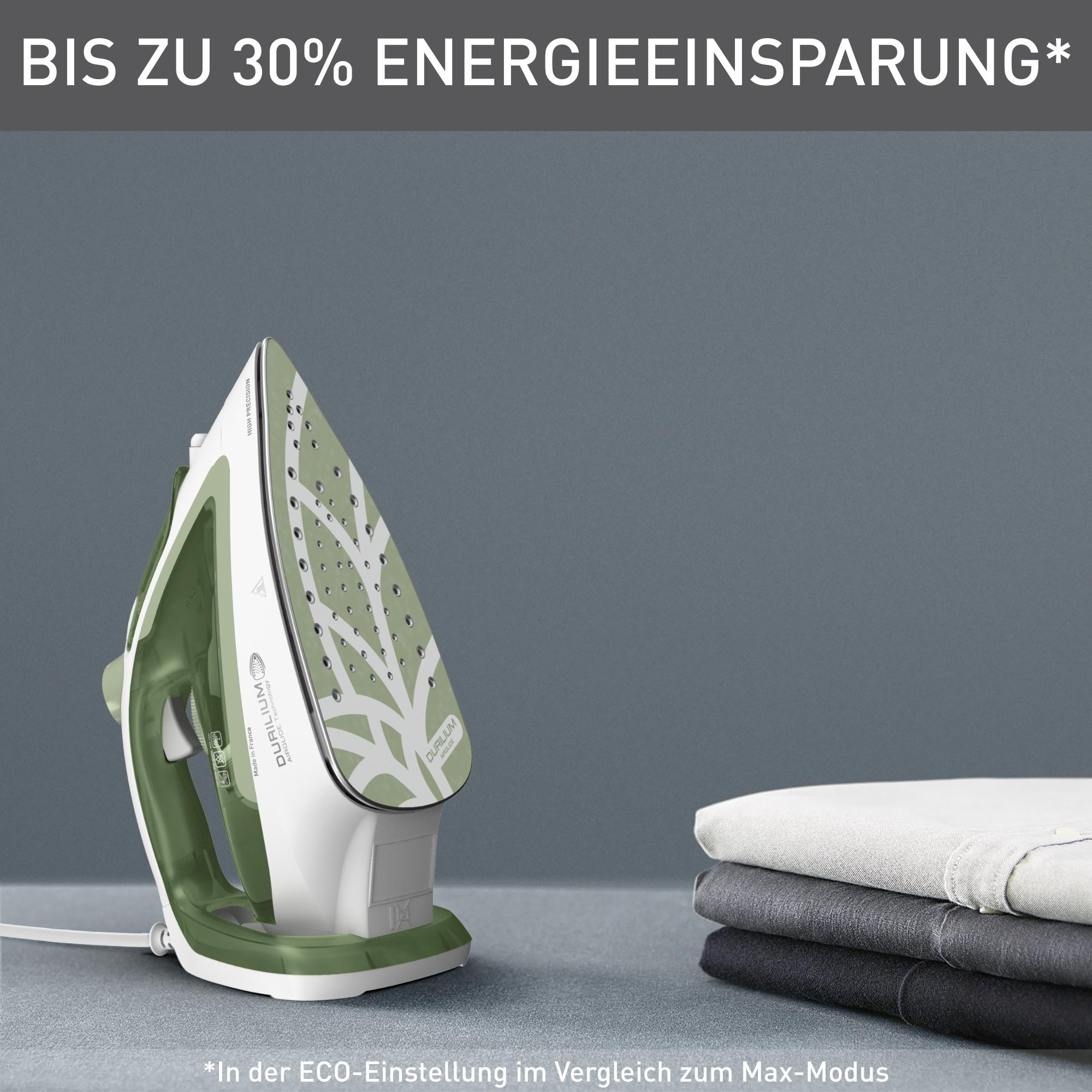 Grünes und weißes Bügeleisen auf grauer Oberfläche. Kleidung ist rechts gestapelt. Text: Energieeinsparung.