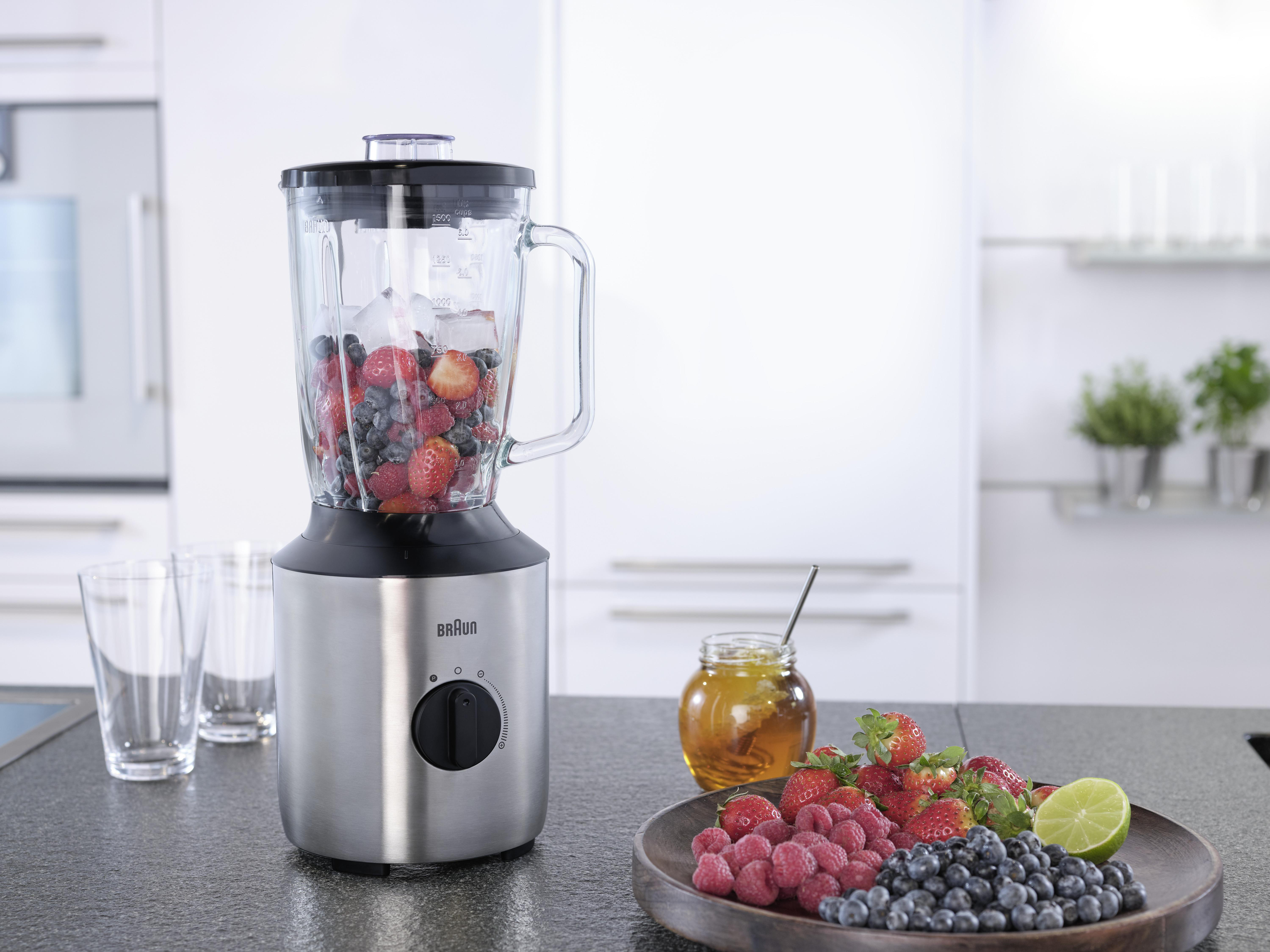 Mixer mit Früchten in einer Küchenzeile. Früchte und Honigglas daneben.