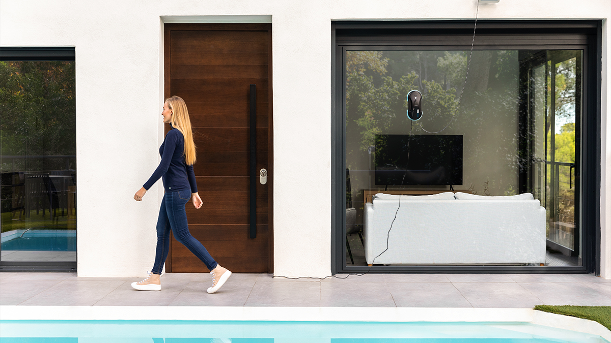 Mujer caminando frente a una casa con una ventana de cristal y un robot limpiacristales.