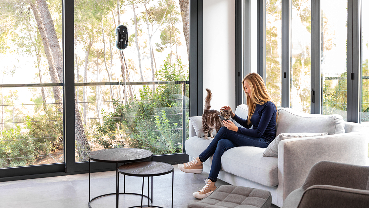 Mujer sentada en un sofá con un gato, robot limpiador en ventana, vista de árboles afuera.