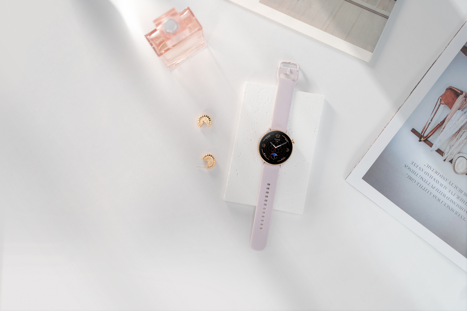Reloj, aretes y perfume sobre una superficie blanca. El reloj tiene una correa rosa.