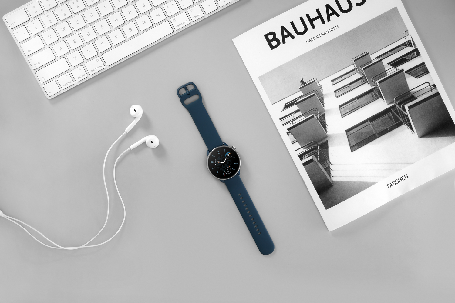 Un reloj inteligente y auriculares en un escritorio gris, junto a un teclado blanco y una revista Bauhaus.