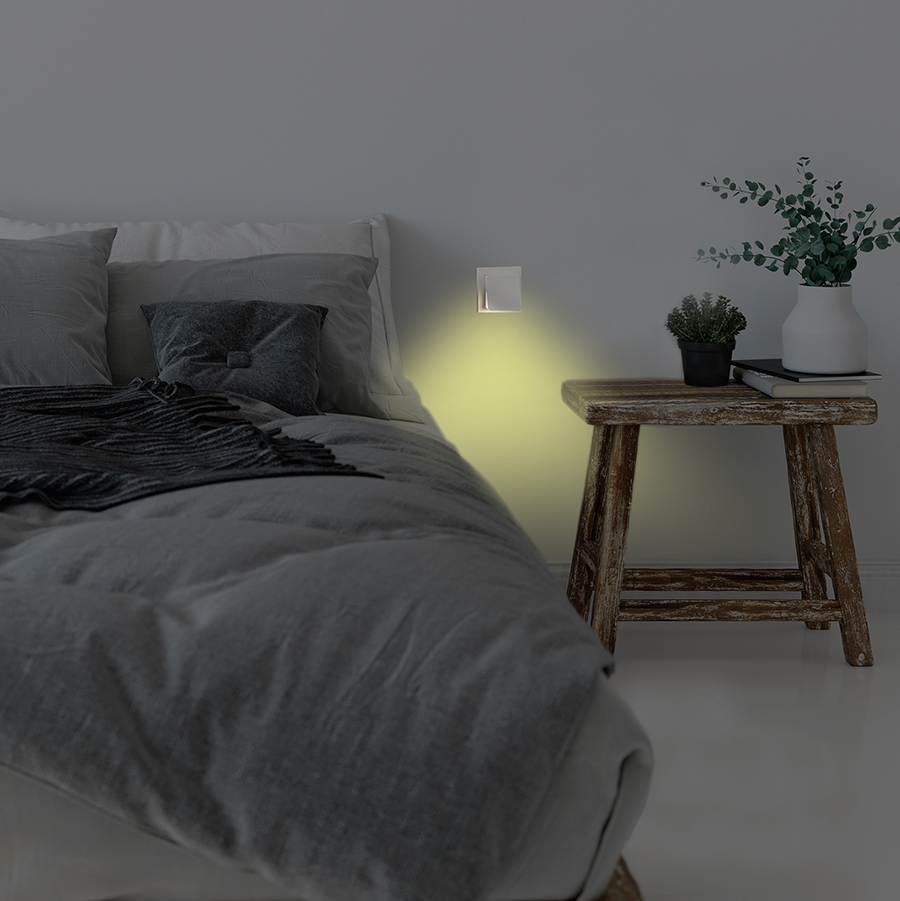 A bedroom with a bed, nightstand, and a wall with a light. The bed is covered in gray bedding.