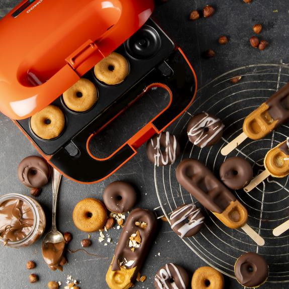 Donut maker with donuts, donut pops, and ingredients on a dark surface. A jar of chocolate is near.