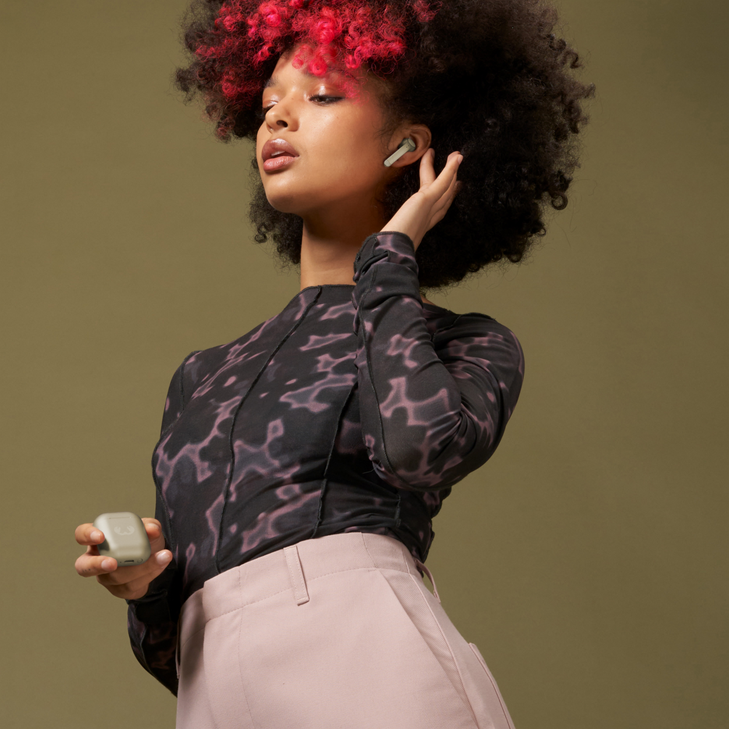 Vrouw met koptelefoon, houdt een etui vast, met roze broek en rood haar.