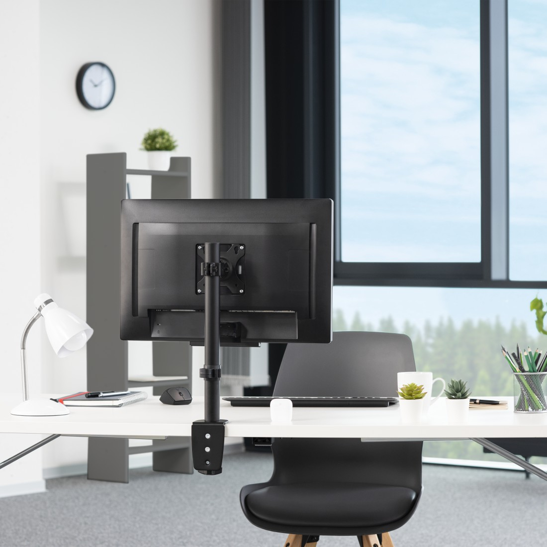 Escritorio con monitor en un soporte, reloj en la pared y ventana con vista.