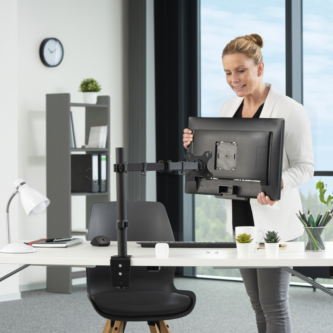 Mujer sostiene un monitor, junto a un brazo de monitor en un escritorio, en una oficina.
