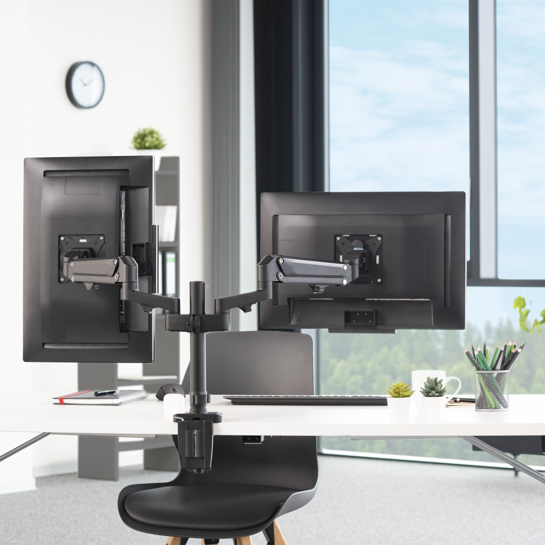 Configuración de doble monitor en escritorio blanco. Un reloj y lápices también están presentes.