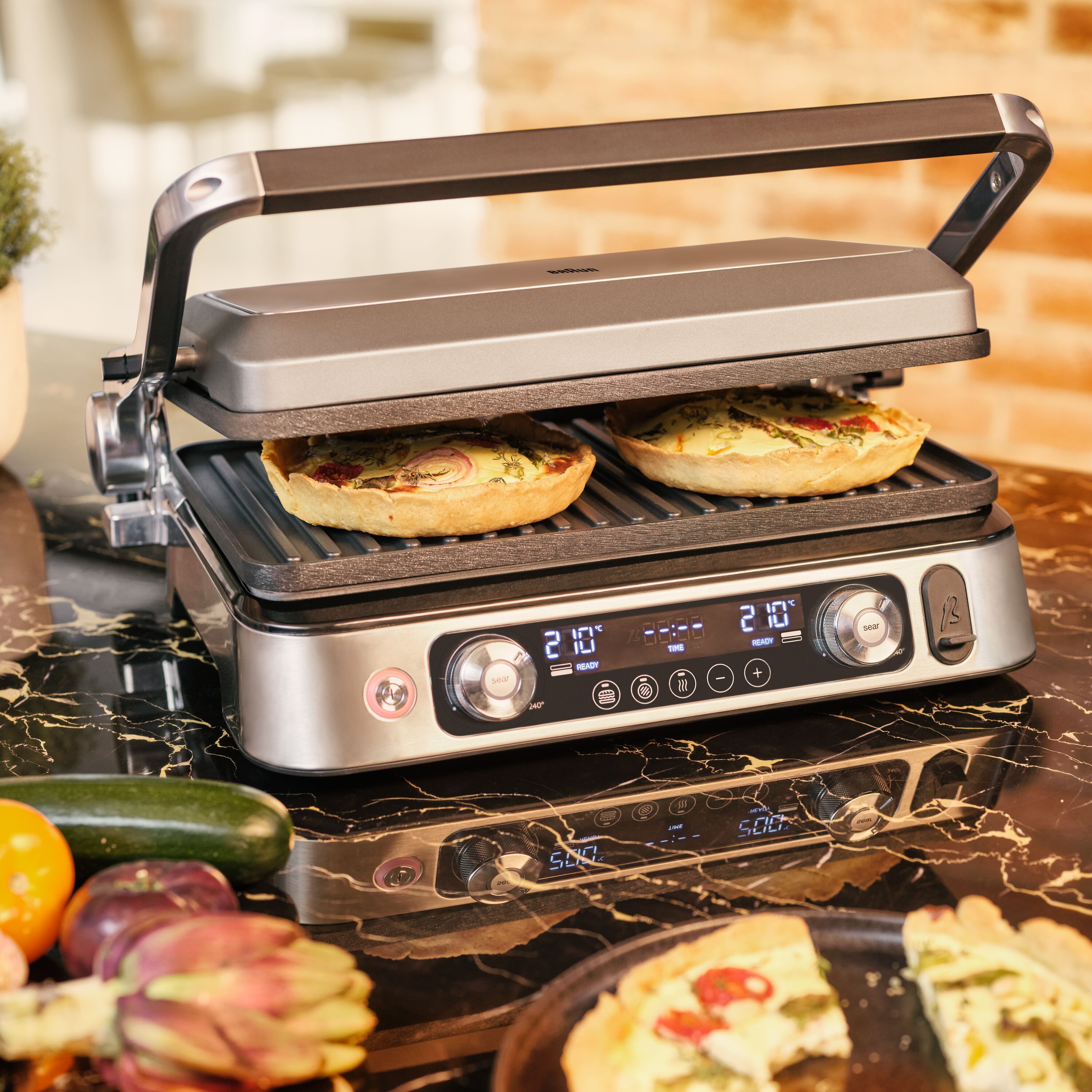 Elektrischer Grill mit zwei Quiches, auf schwarzer Marmorarbeitsplatte, mit anderen Lebensmitteln.