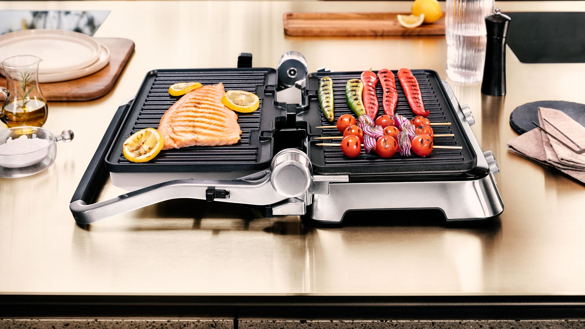 Offener Grill mit Lachs, Zitronenscheiben, Tomaten und Spießen. Der Grill steht auf glänzender Oberfläche.