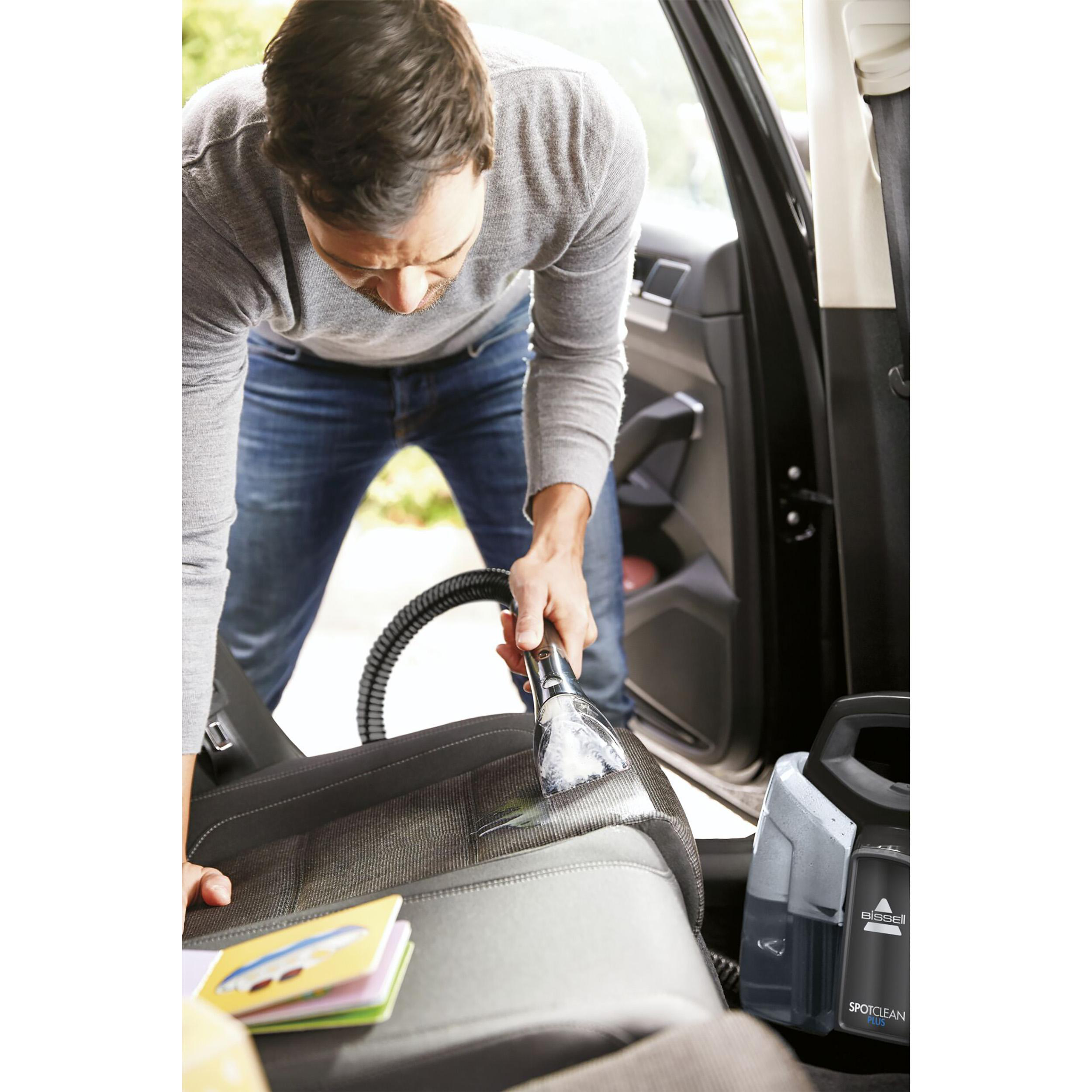 Mann reinigt Autositz mit Bissell SpotClean, Bücher und geöffnete Autotür sichtbar.