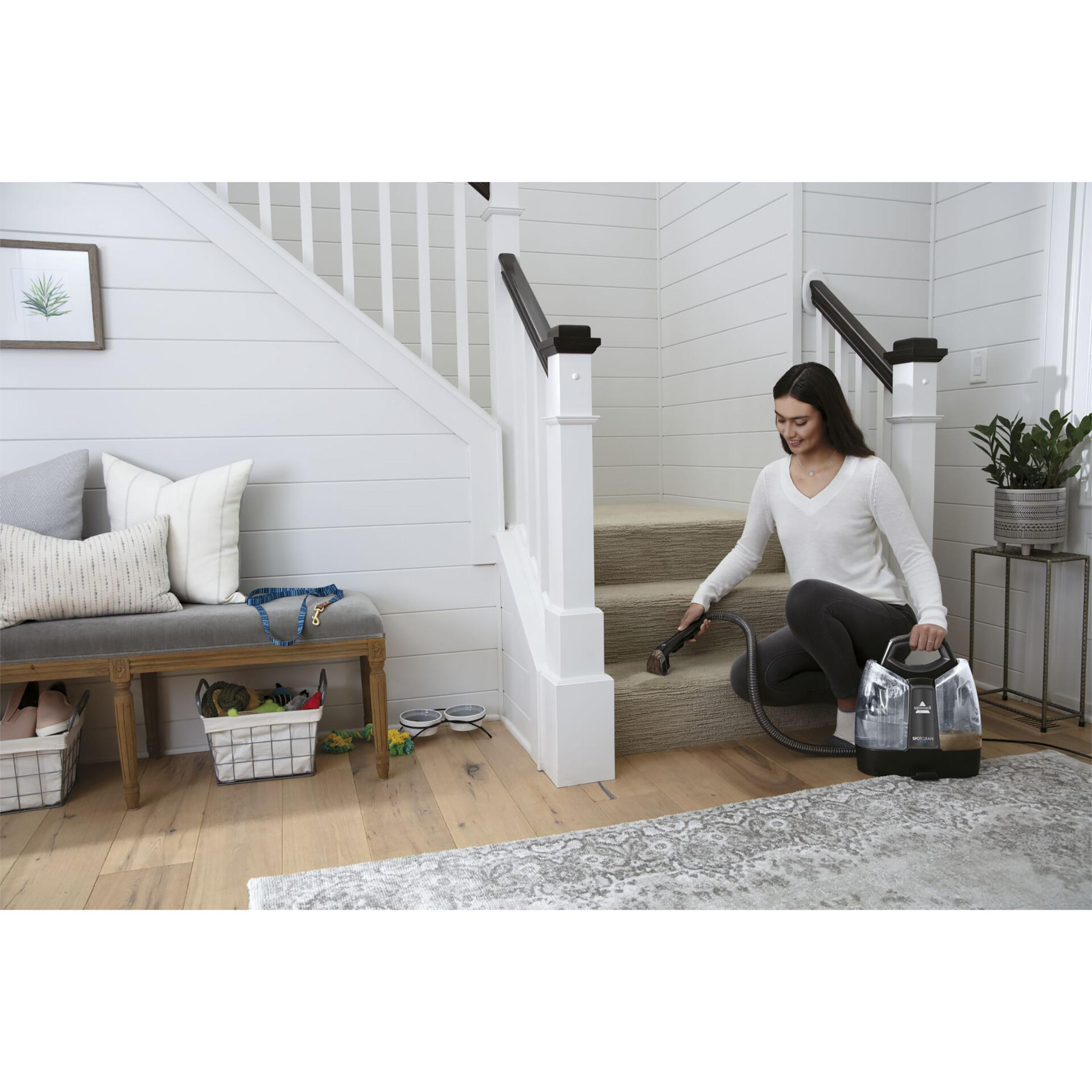 Eine Frau reinigt eine Teppichtreppe mit einem Bissell-Reiniger. Eine Bank und Treppe sind im Hintergrund.