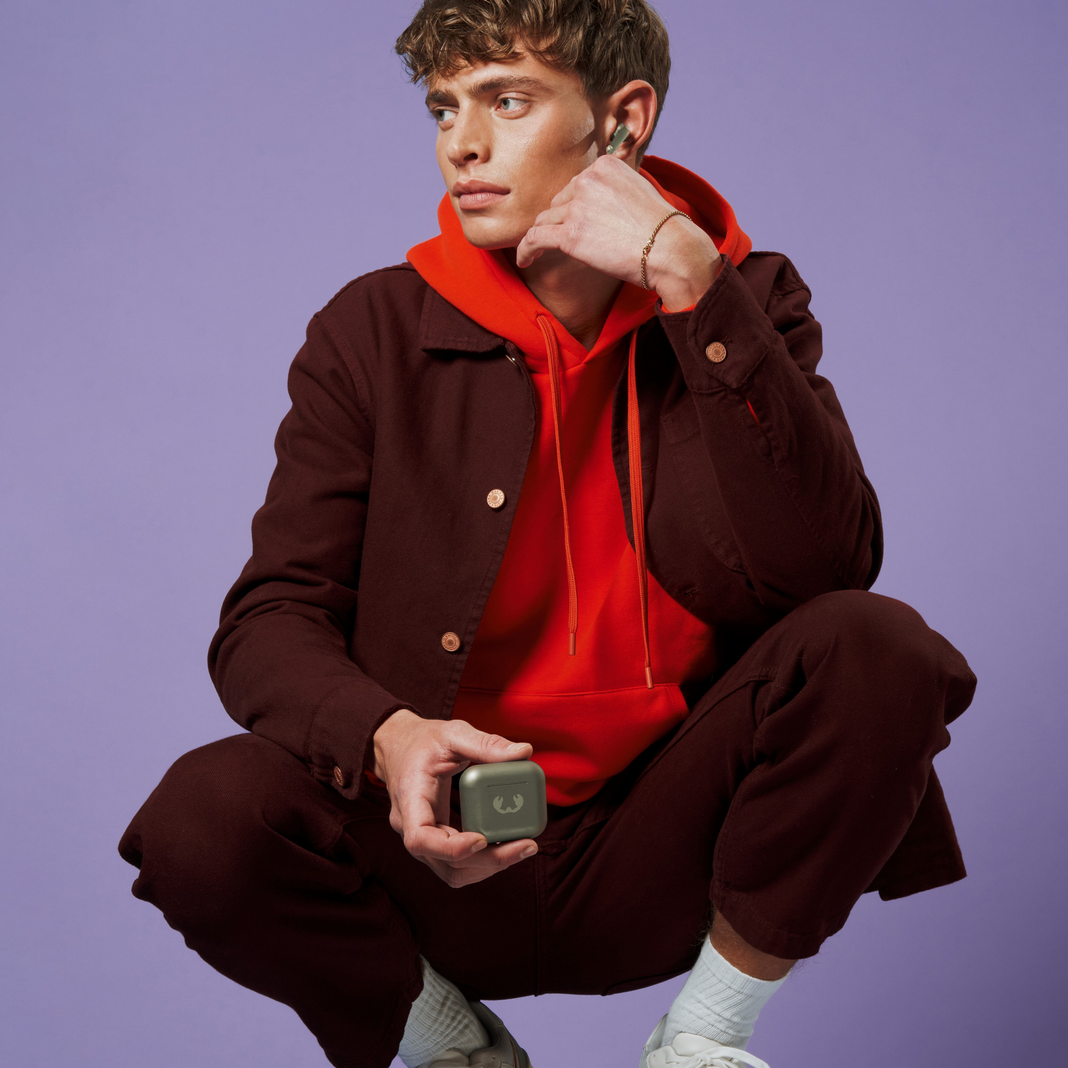 Een man die een hoesje met oortjes vasthoudt. Hij draagt een jas, hoodie en broek, op een paarse achtergrond.
