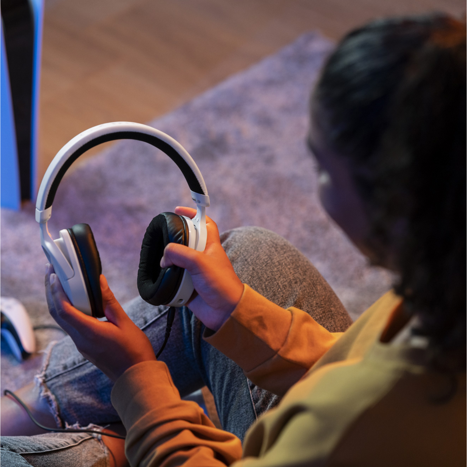 Persoon houdt witte koptelefoon vast. Zwarte oorschelpen. Grijze vloerkleed en PS5 op de achtergrond.