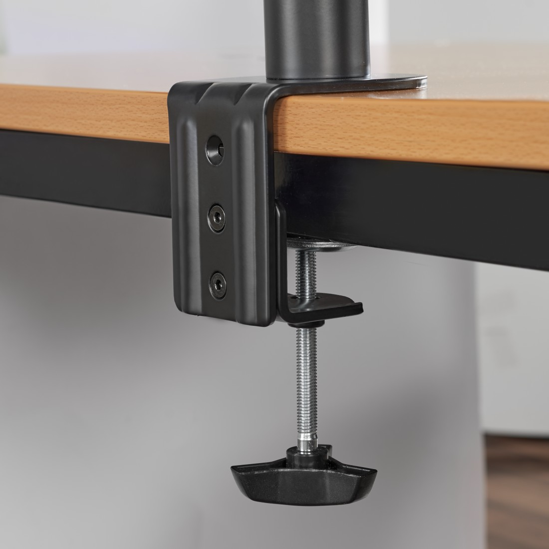 Abrazadera de escritorio negra con tornillo. Montada en mesa de madera con fondo gris claro.