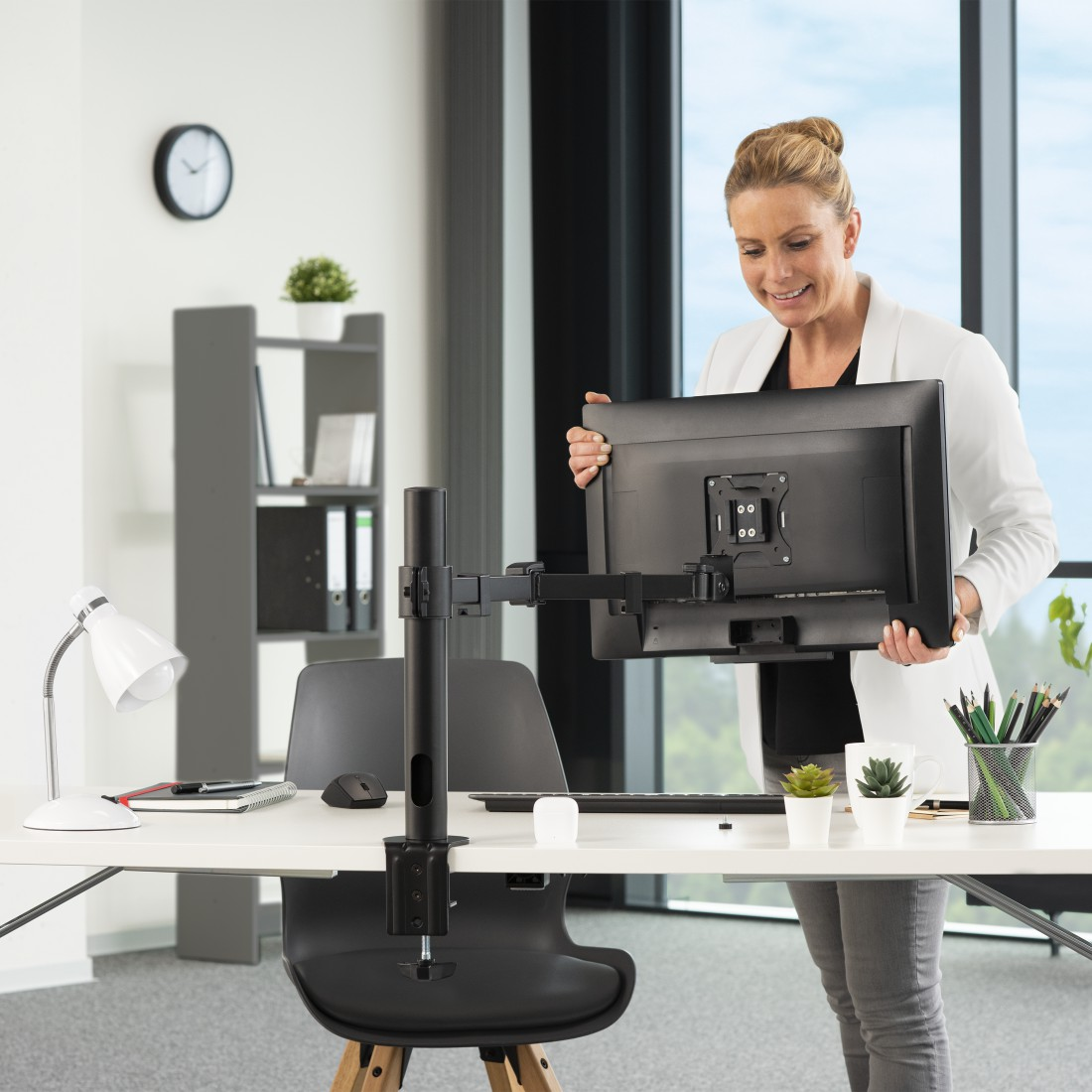 Mujer sosteniendo un monitor, cerca de escritorio con brazo. Ambiente de oficina.
