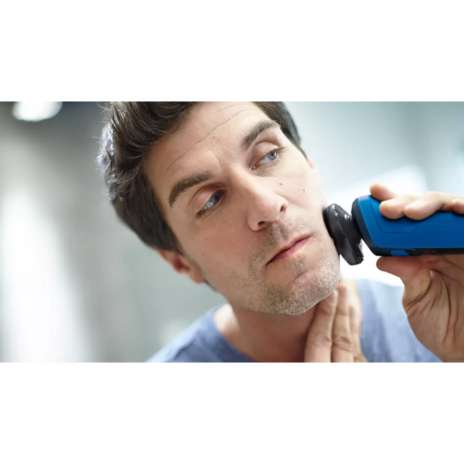 A man shaves his face with a blue and black electric razor in a bathroom.