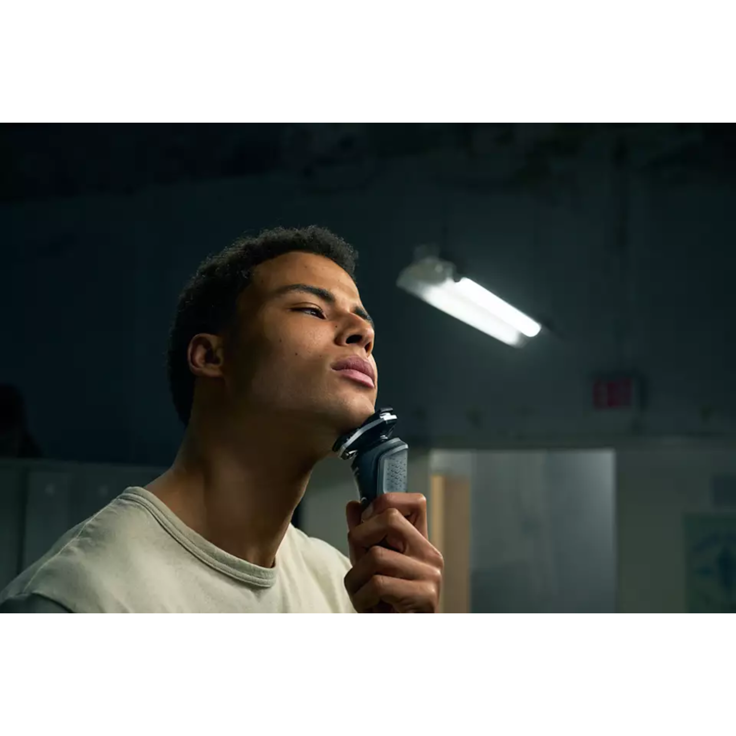 Man shaves with an electric razor. The background is dark with a long light.