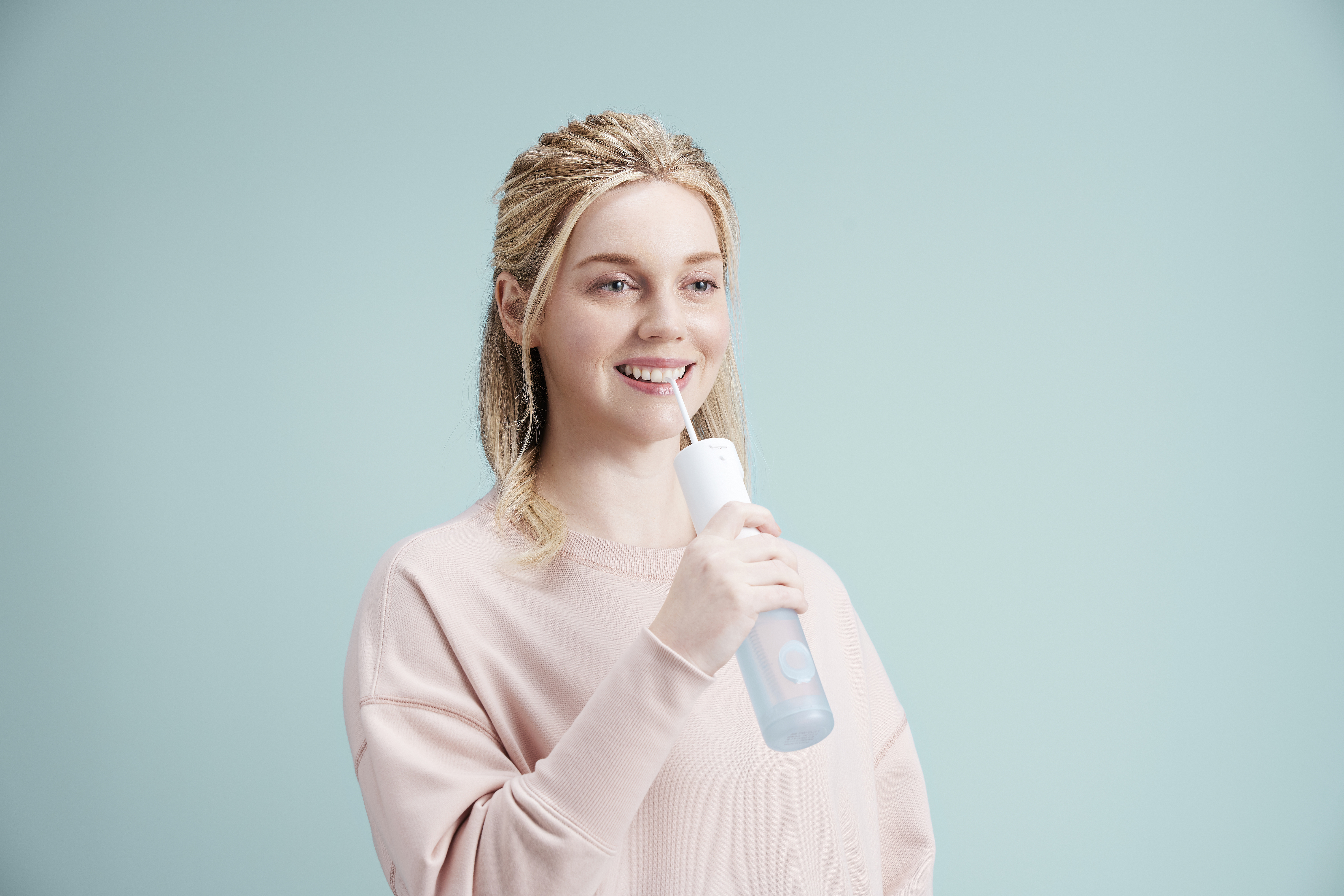 Una mujer con cabello rubio usa un irrigador bucal. Está sonriendo, con un fondo azul.