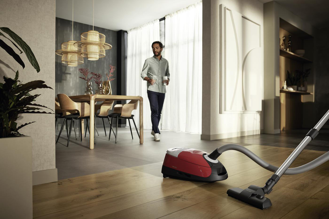 Ein Mann geht in einem modernen Wohnzimmer mit einem Staubsauger im Vordergrund, Esstisch und Pflanze im Hintergrund.