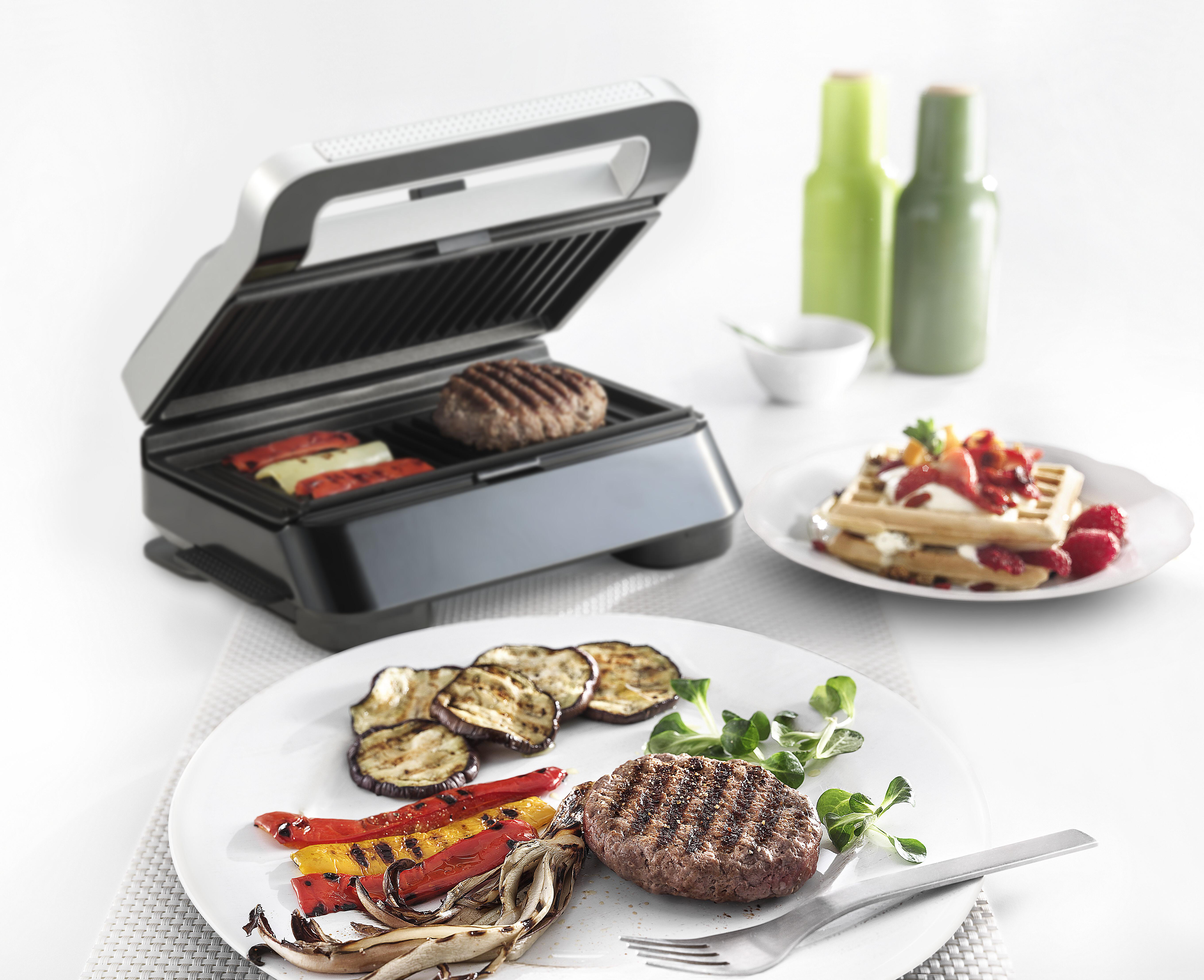 Gegrilltes Essen auf weißen Tellern, Waffel mit Obst, Grill und zwei Soßenflaschen auf weißer Oberfläche.