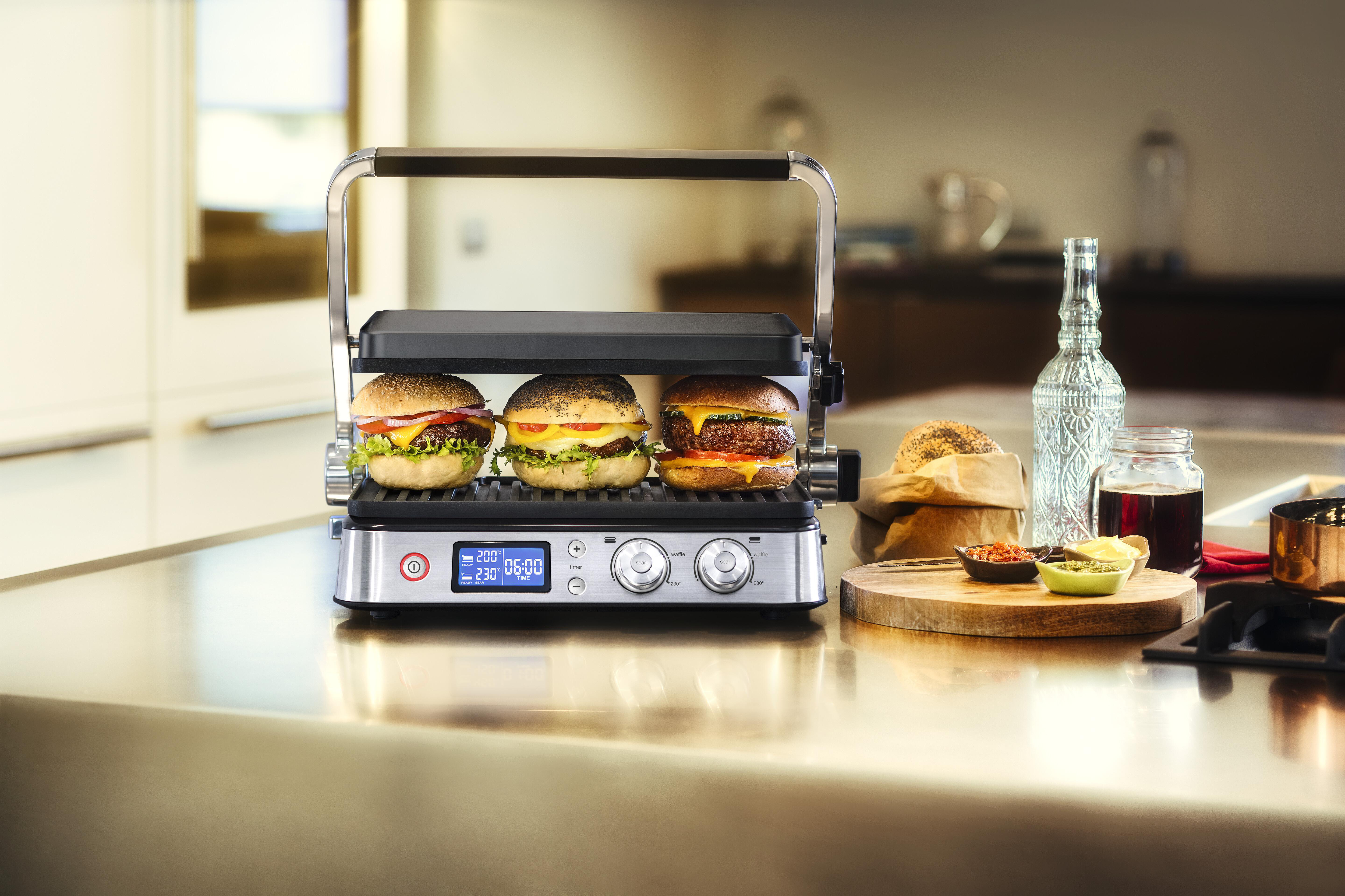 Ein silberner Grill mit drei Burgern, plus Gewürzen, auf einer Küchentheke, glänzend.