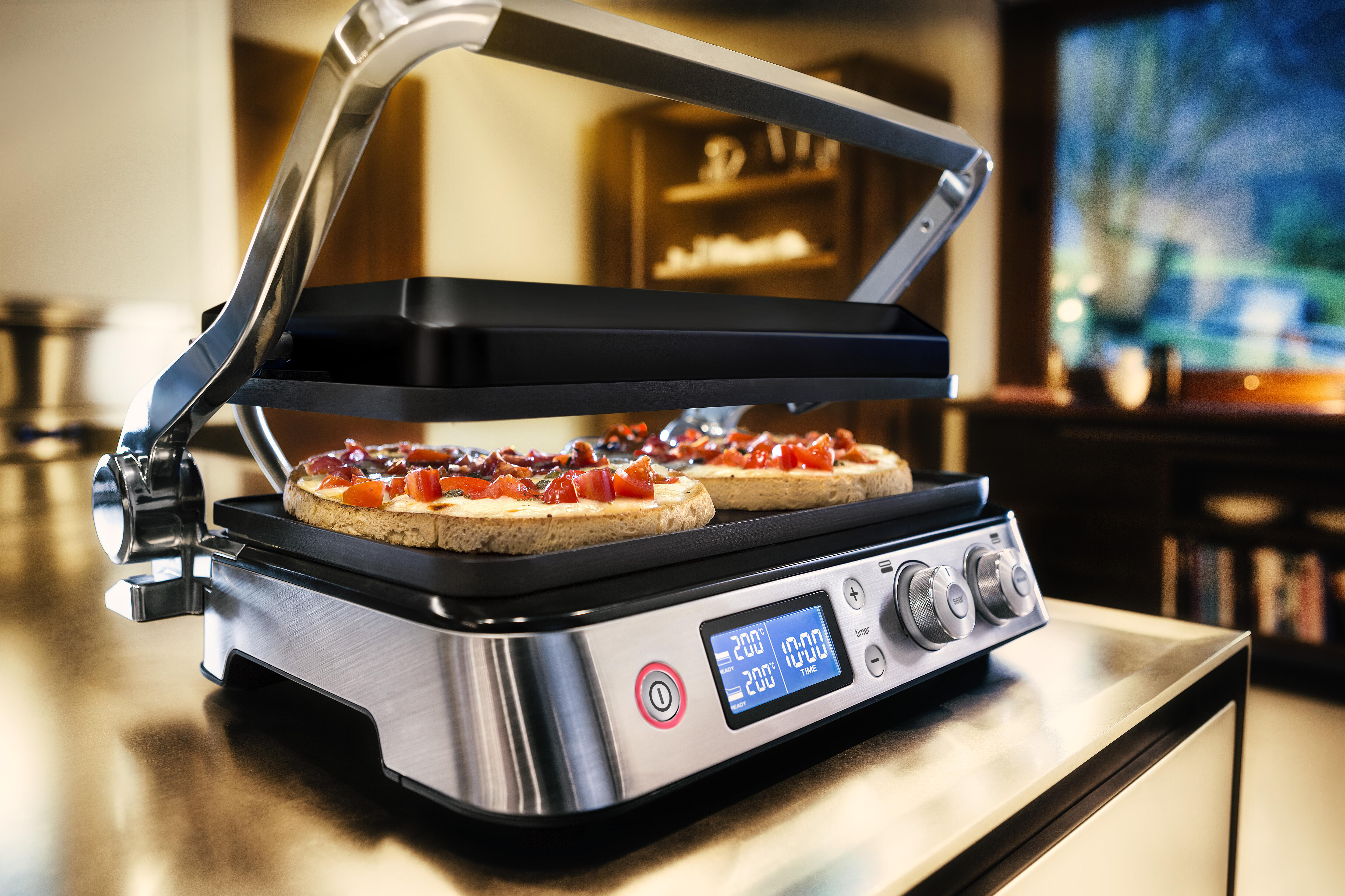 Eine silberne und schwarze Panini-Presse mit zwei Sandwiches, die auf einer Edelstahl-Arbeitsplatte garen.