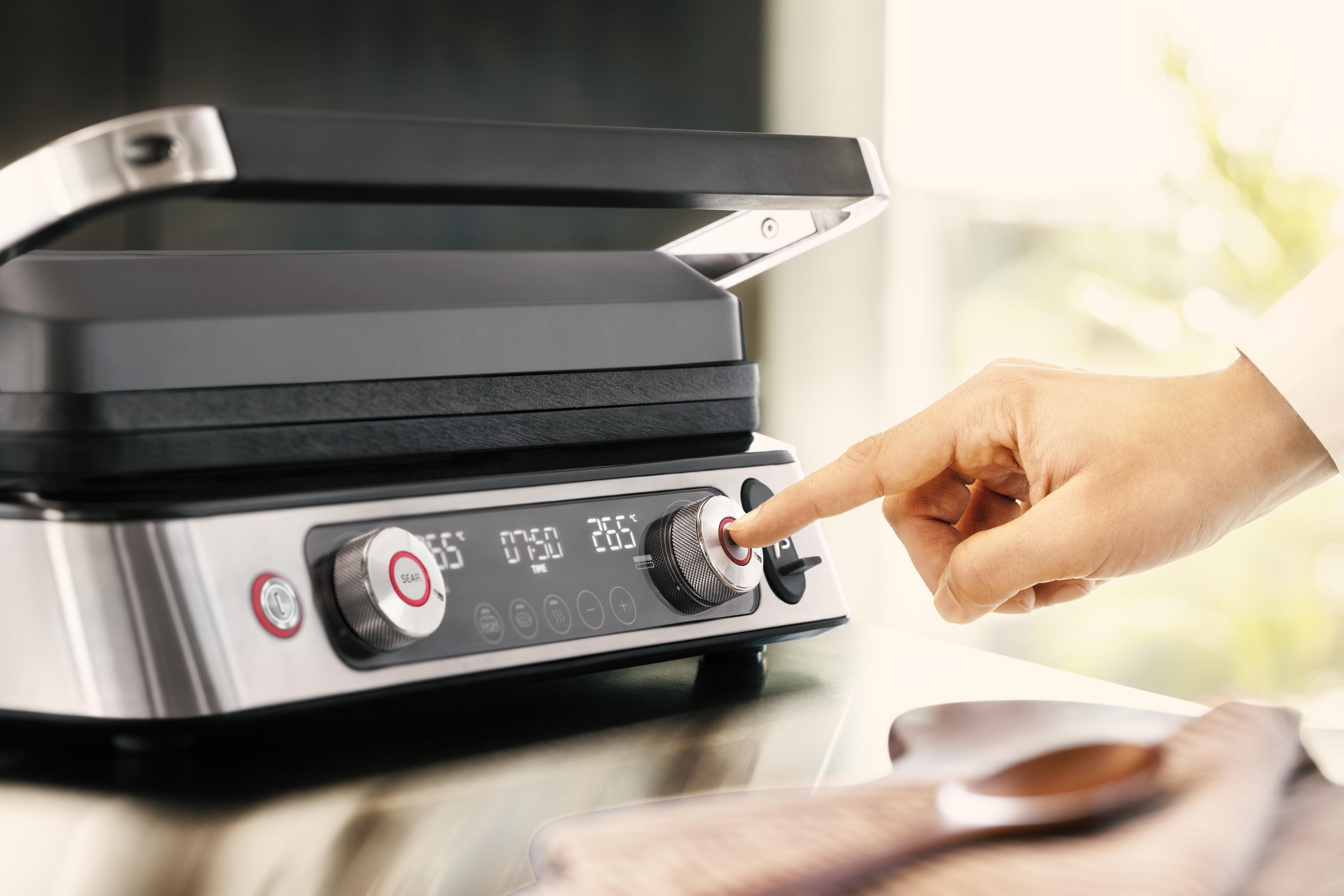 Eine Hand drückt eine Taste auf einem silbernen Grill. Ein Timer zeigt die Temperatur. Verschwommener Hintergrund.